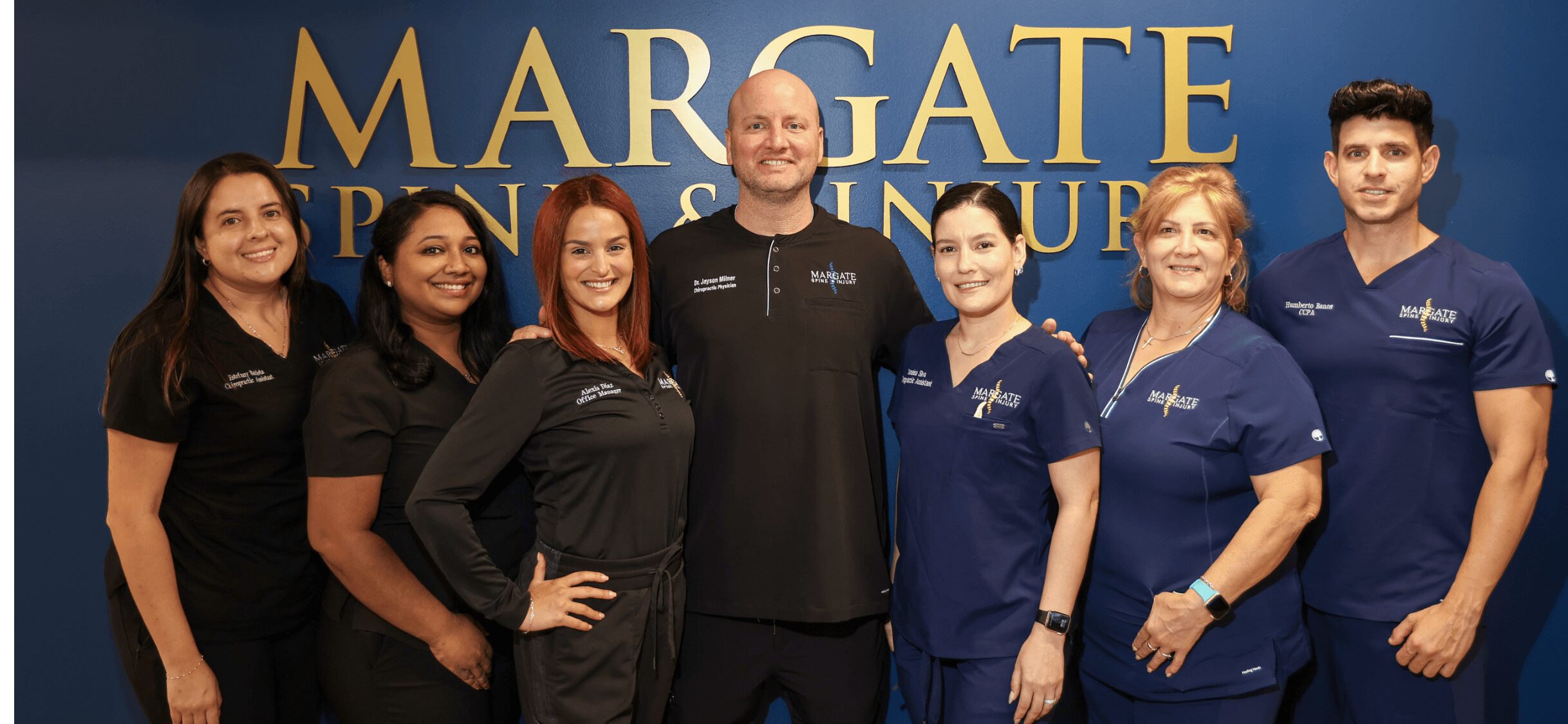 Group of seven people standing in front of a blue background with gold text, smiling at the camera.