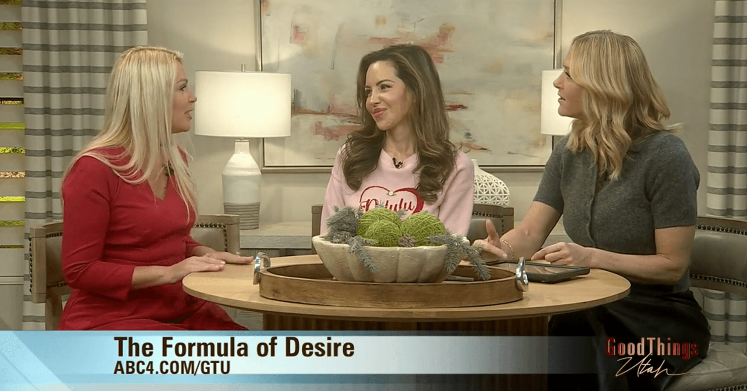 Three women sit around a table with a bowl of vegetables, engaged in conversation in a living room setting.