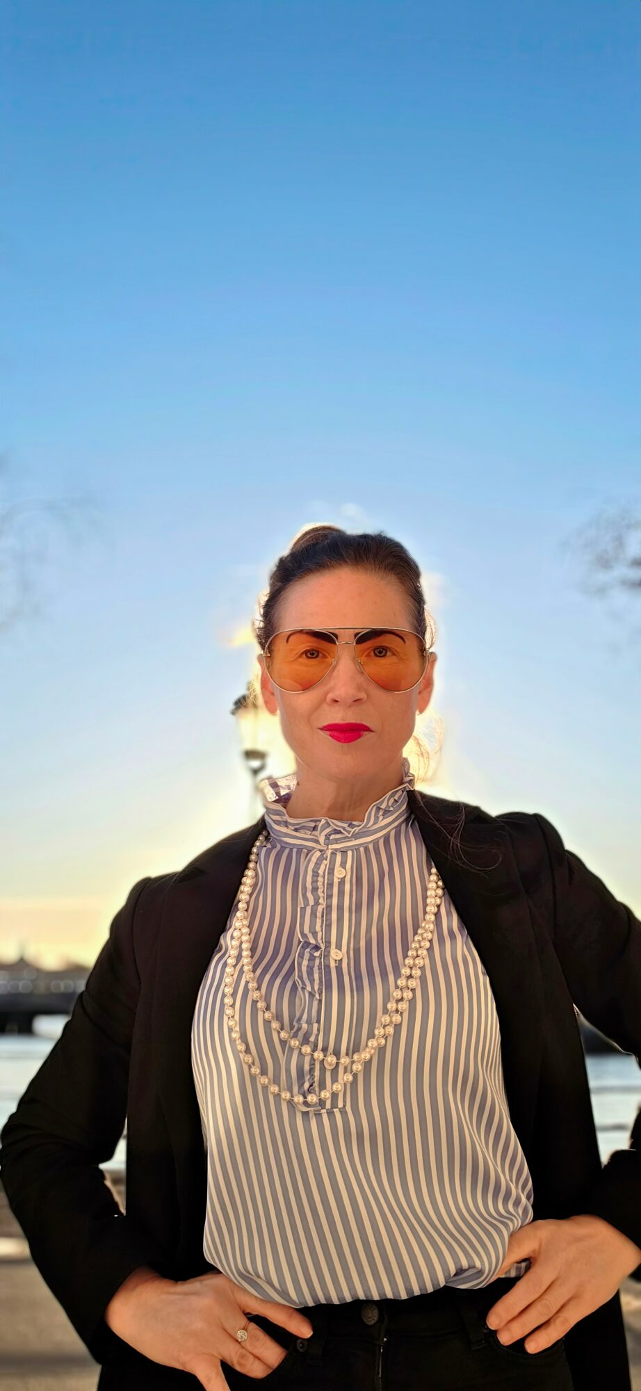 Woman with glasses and red lipstick standing outdoors against a blue sky, wearing a striped blouse and black jacket.