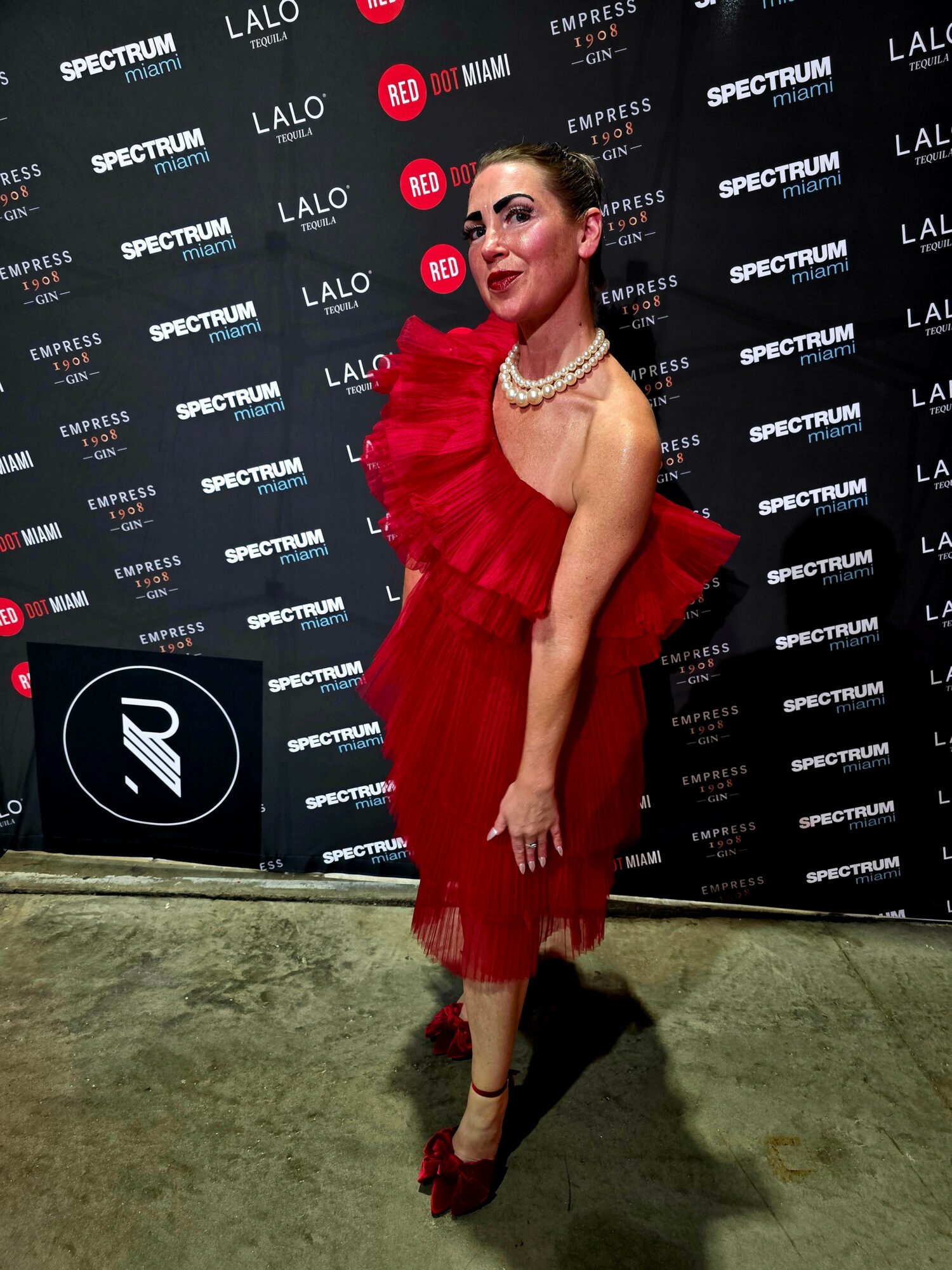 Woman in red dress with ruffles, pearl necklace, and red shoes, posing in front of a black backdrop with logos.