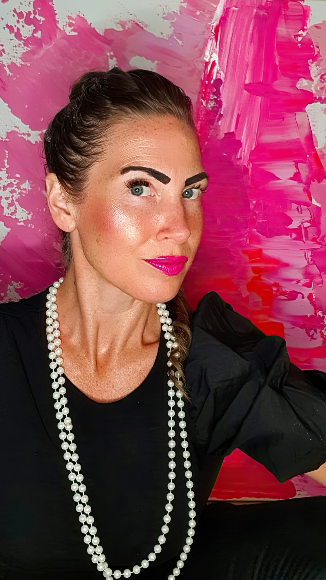 Woman with dark hair in a bun, wearing pearl necklace and black top, against pink and purple abstract background.