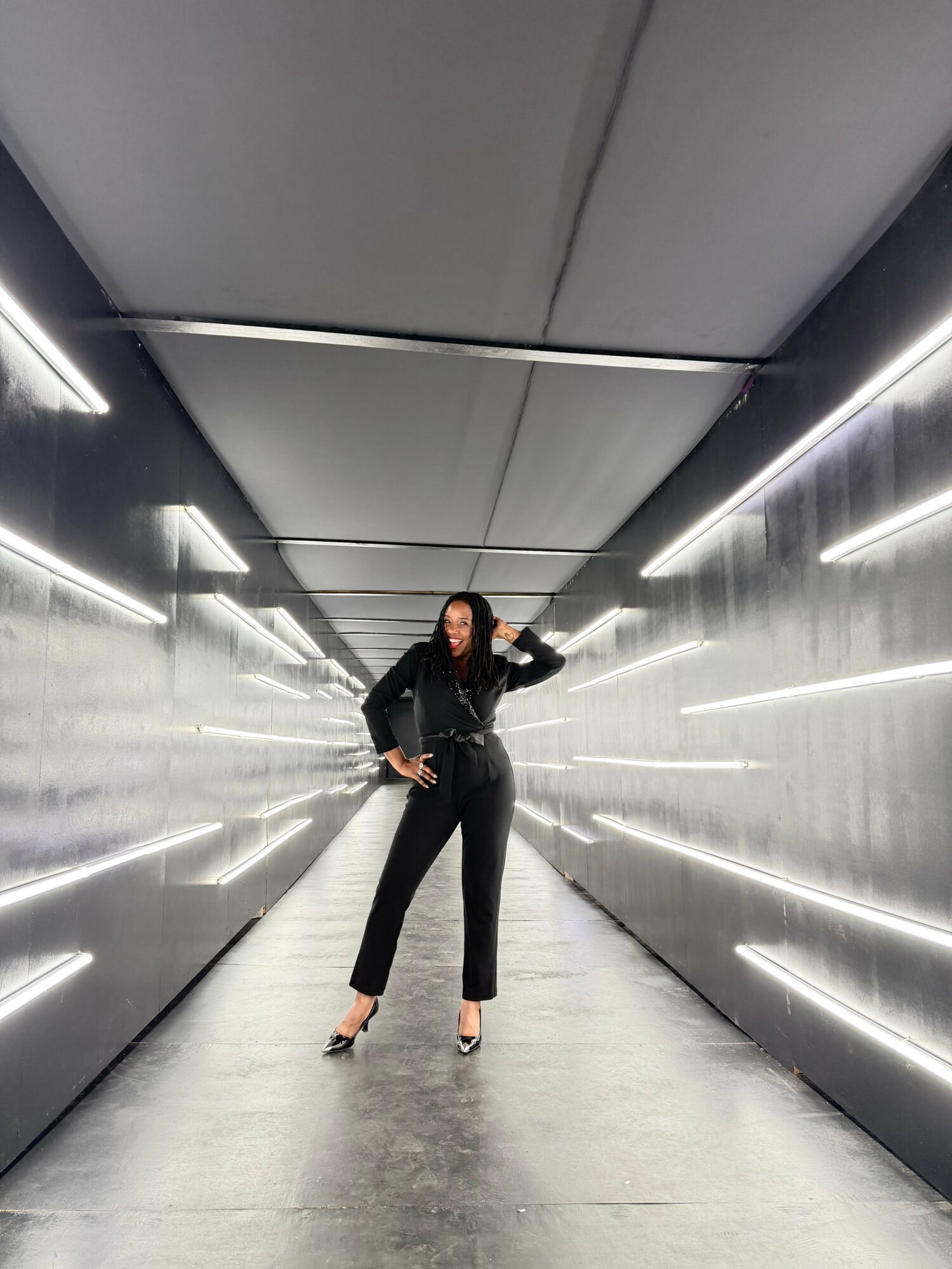 Woman standing in a futuristic corridor with illuminated walls and ceiling, hands on hips, wearing black outfit and heels.