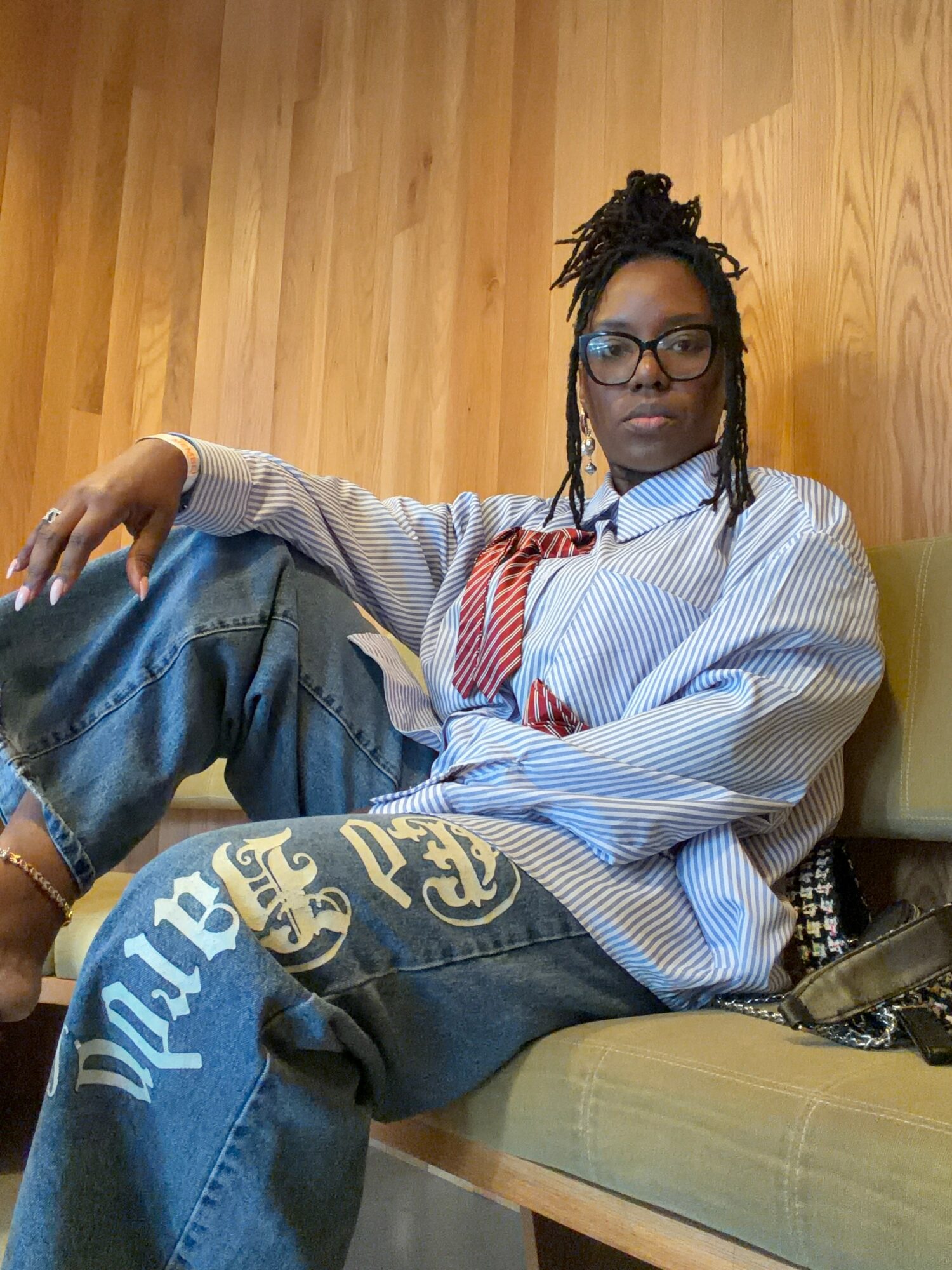 Person with glasses and braided hair sitting on a bench with wooden wall background, wearing a striped shirt and baggy jeans.