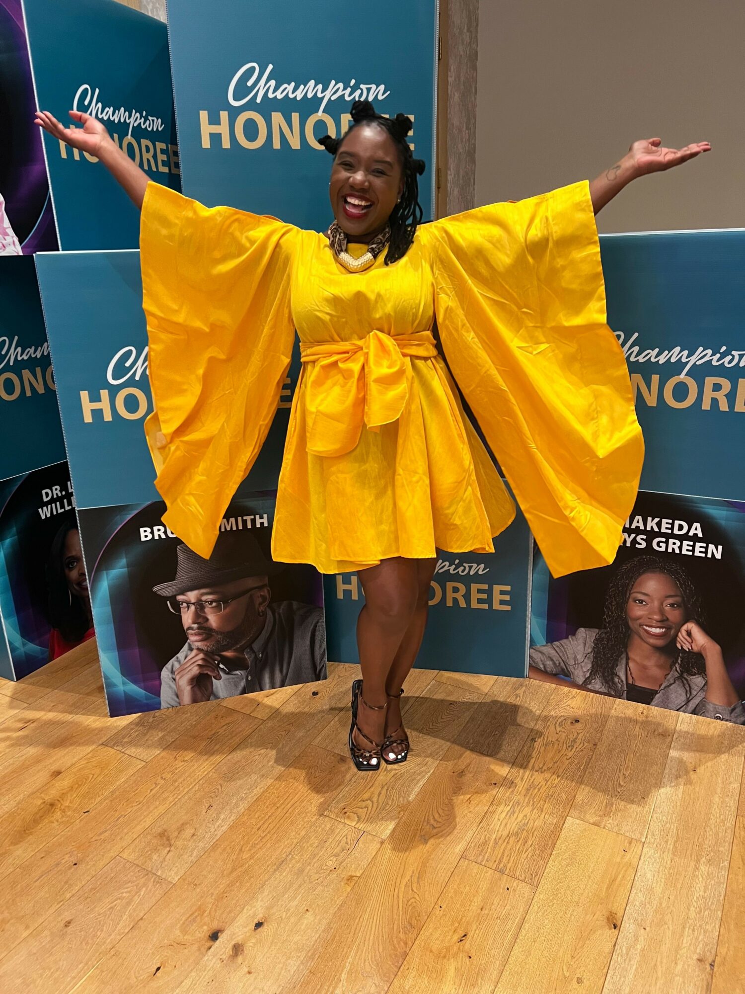 Woman in bright yellow dress with wide sleeves, arms outstretched, smiling, standing on wooden floor in front of blue and purple banners.