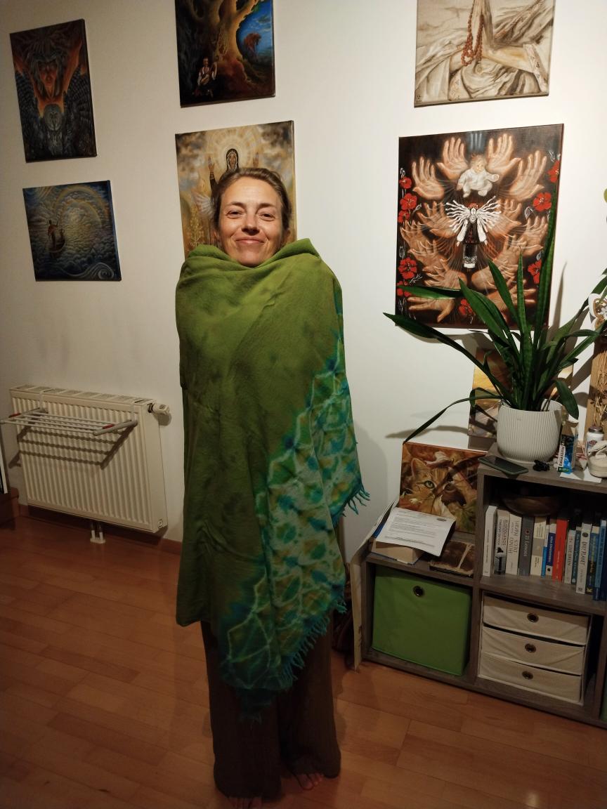Woman standing in art gallery with paintings on wall, green shawl, and a plant on nearby table.