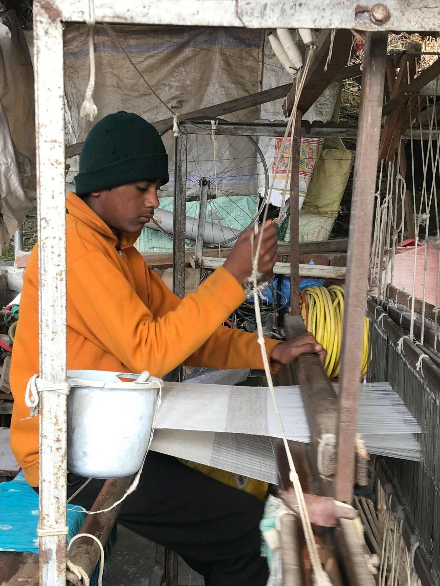 Young person wearing a black beanie and orange jacket working with a loom or weaving machine outdoors.