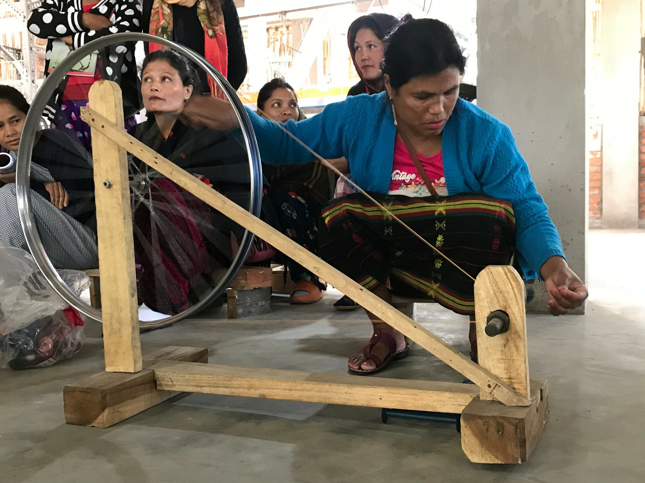 Woman operating a wooden spinning wheel with children watching indoors.