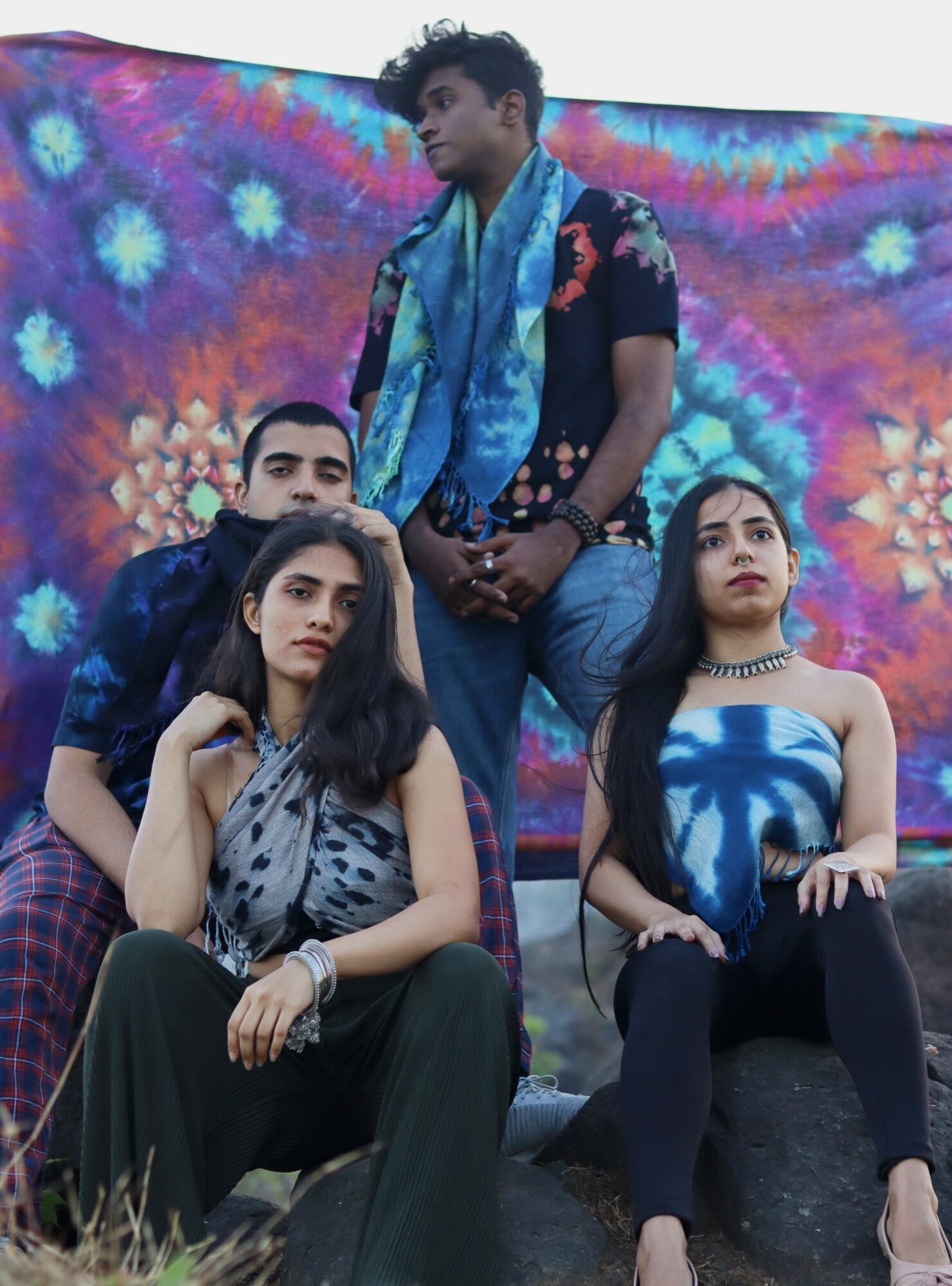 Four young people pose outdoors against a colorful tie-dye backdrop, with two sitting and two standing, dressed in casual clothing.
