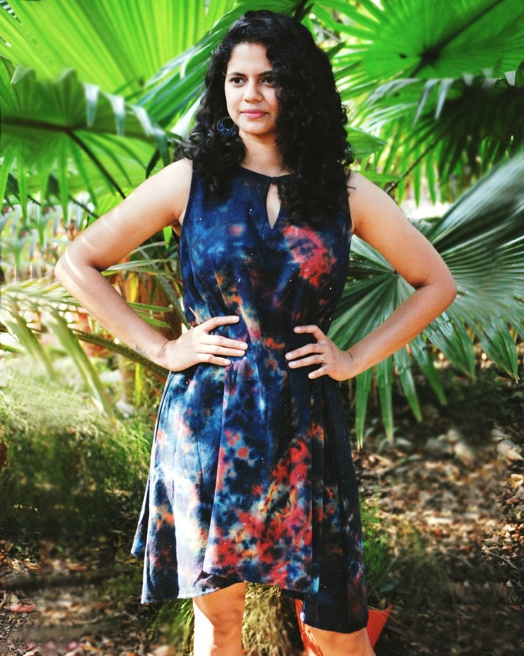 Woman with curly hair in a colorful dress standing outdoors among green plants, hands on hips, smiling.