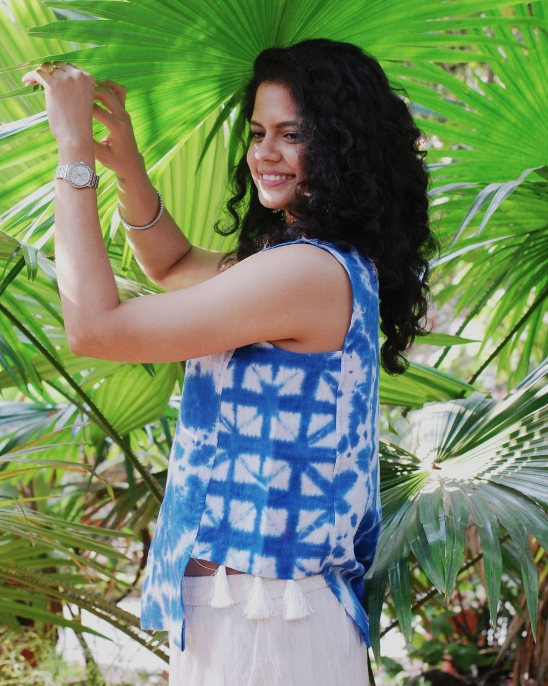Woman with curly hair smiling, wearing a blue and white patterned sleeveless top, standing among green tropical leaves.