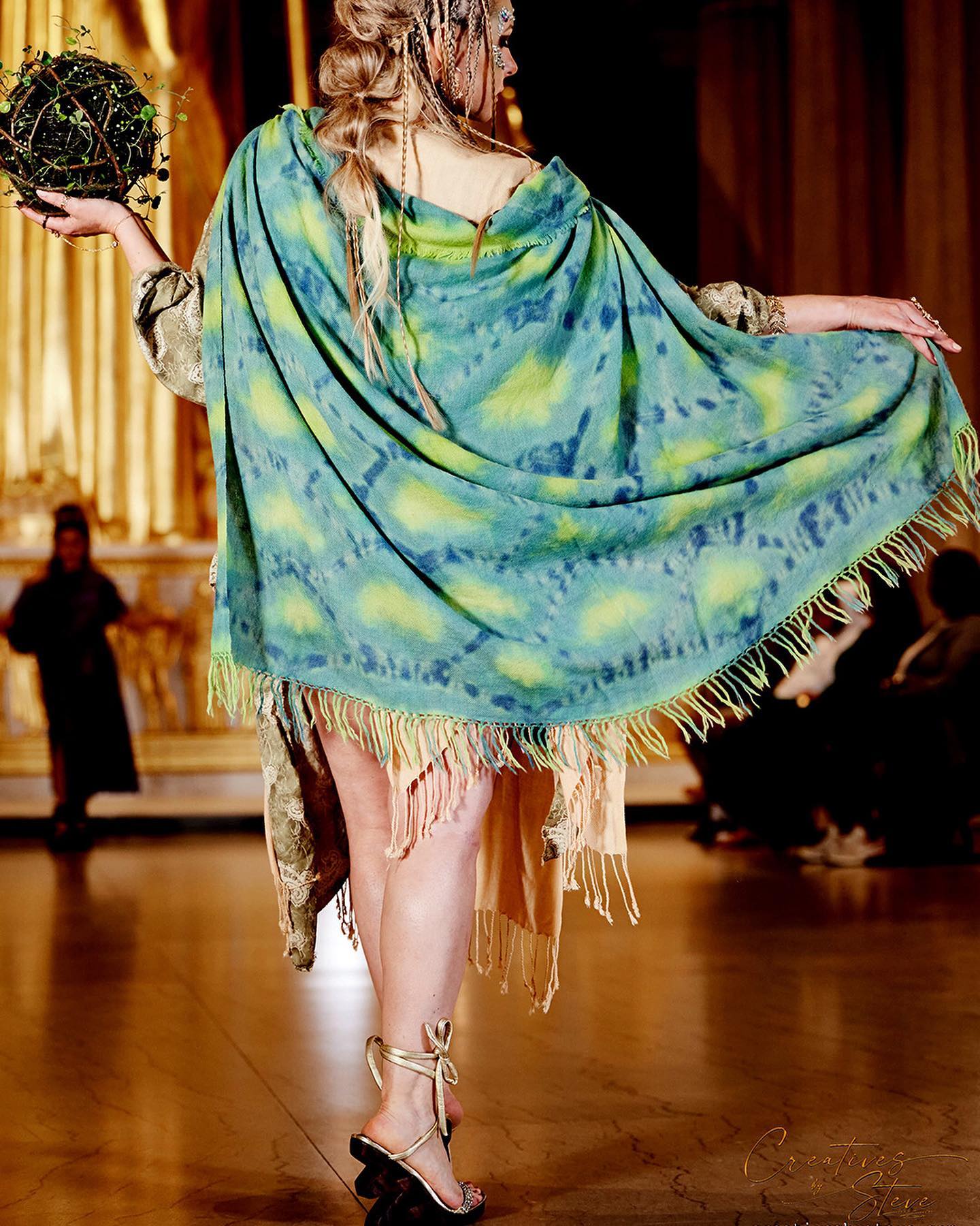 Person on stage holding a globe, wearing a colorful shawl and fringed dress, with a wooden floor and audience in background.