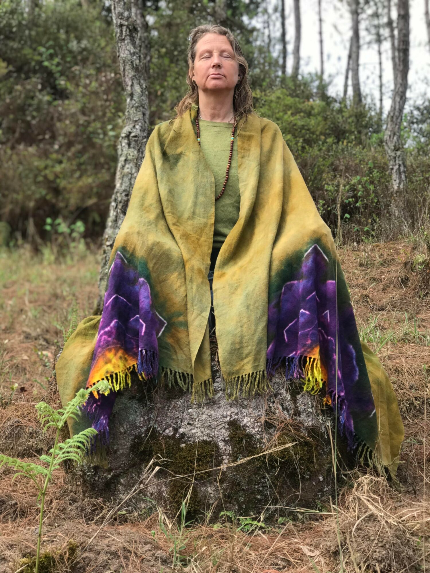 Woman standing outdoors with eyes closed, wearing a green top and a large yellow and purple shawl, surrounded by trees.