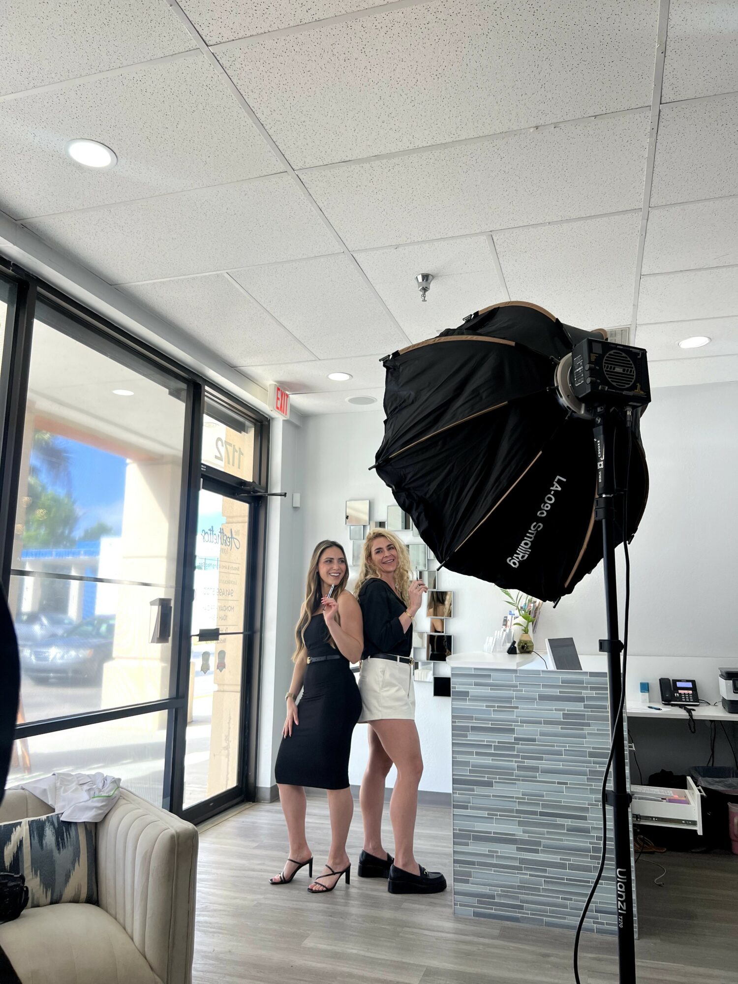 Two women stand inside a room near a large photography umbrella and a counter, smiling at the camera.