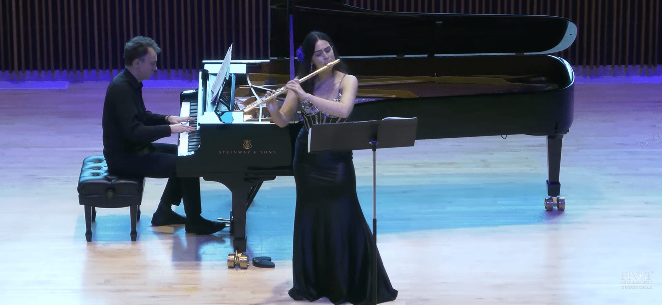 Piano player and flutist perform on stage with a grand piano and purple curtain backdrop.