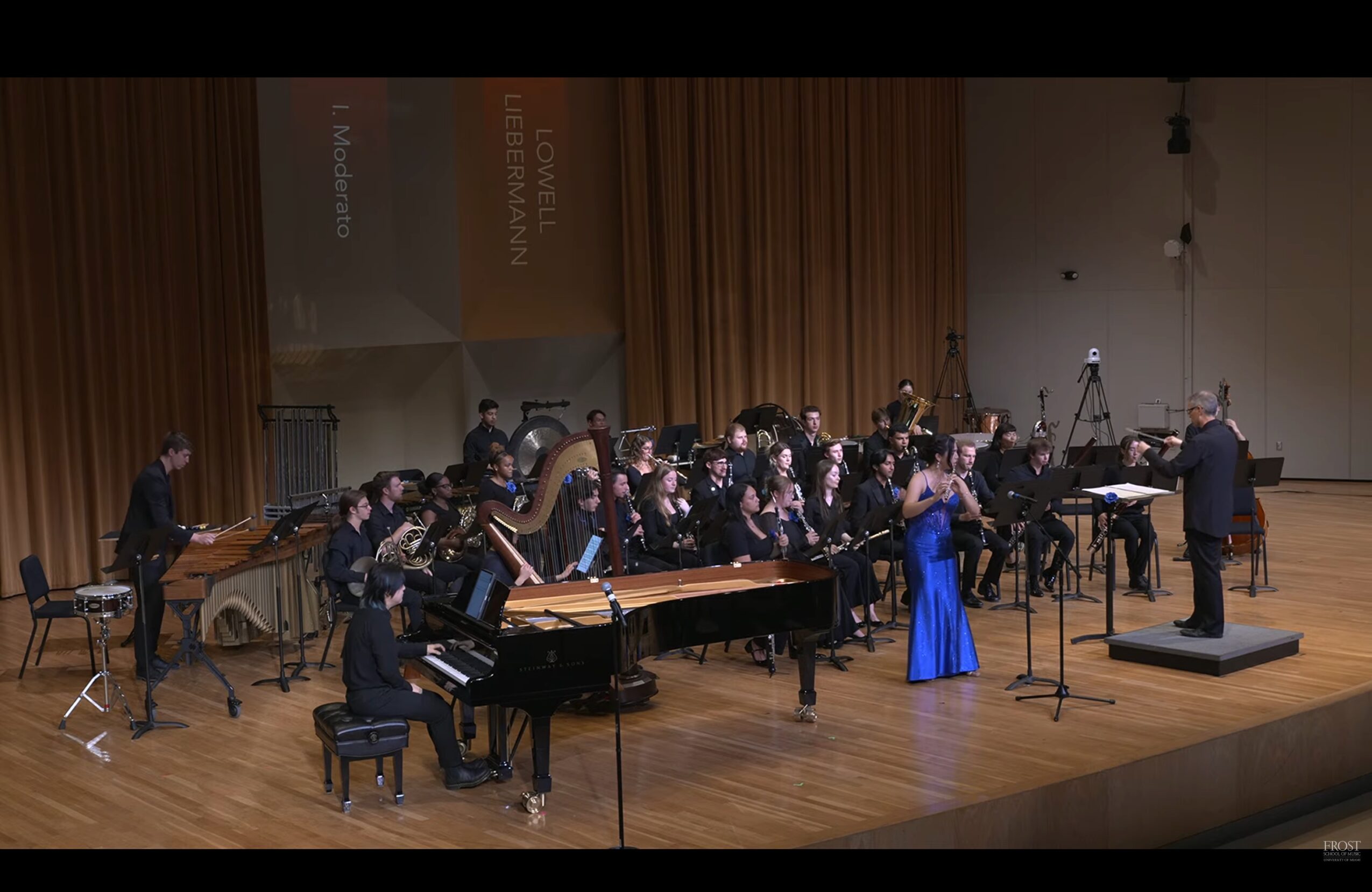Orchestra performing on stage with conductor, pianist, and a singer in a blue dress, in a concert hall.