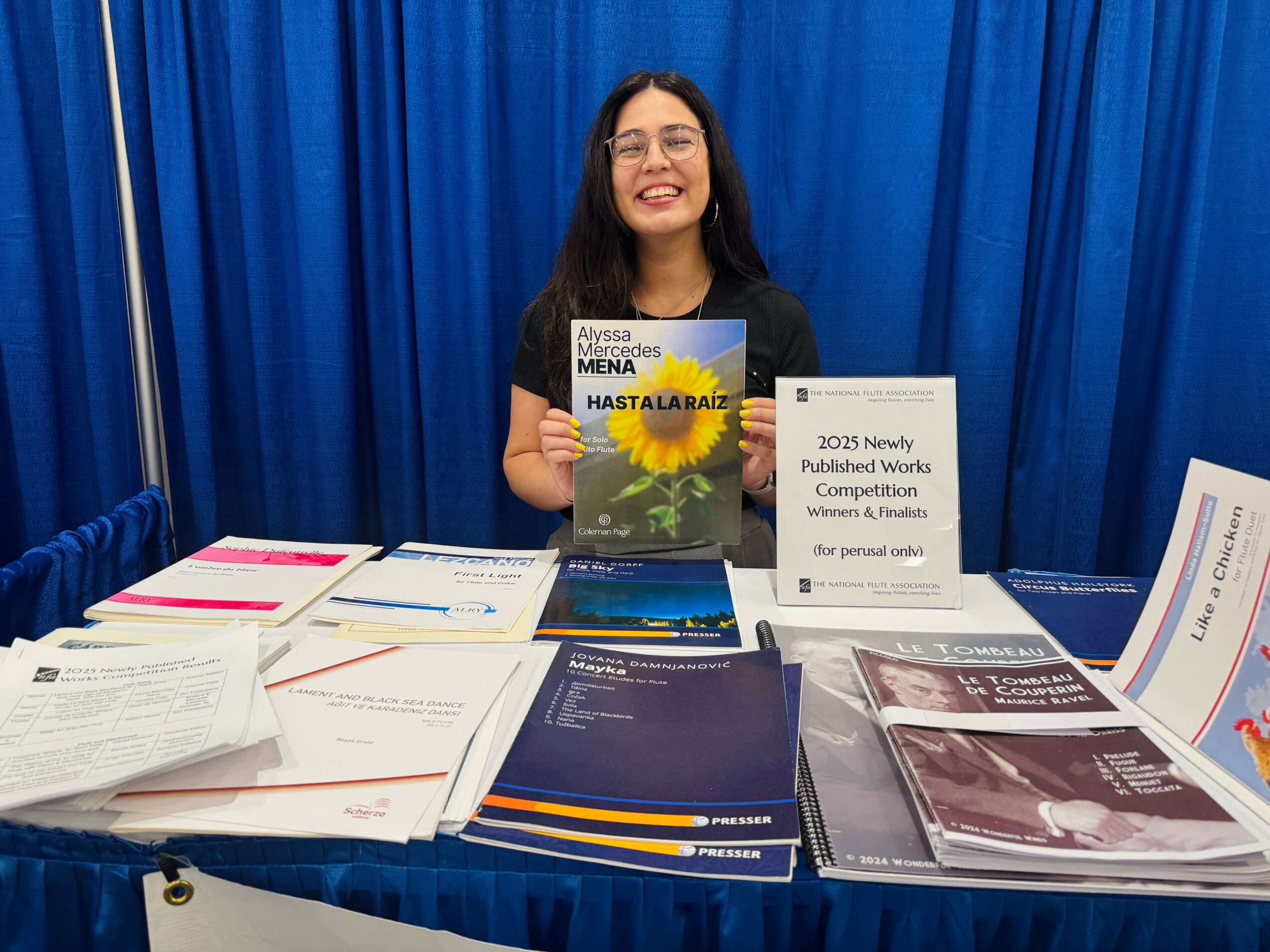 Young woman with glasses smiling behind a table with books and papers, blue curtain background.