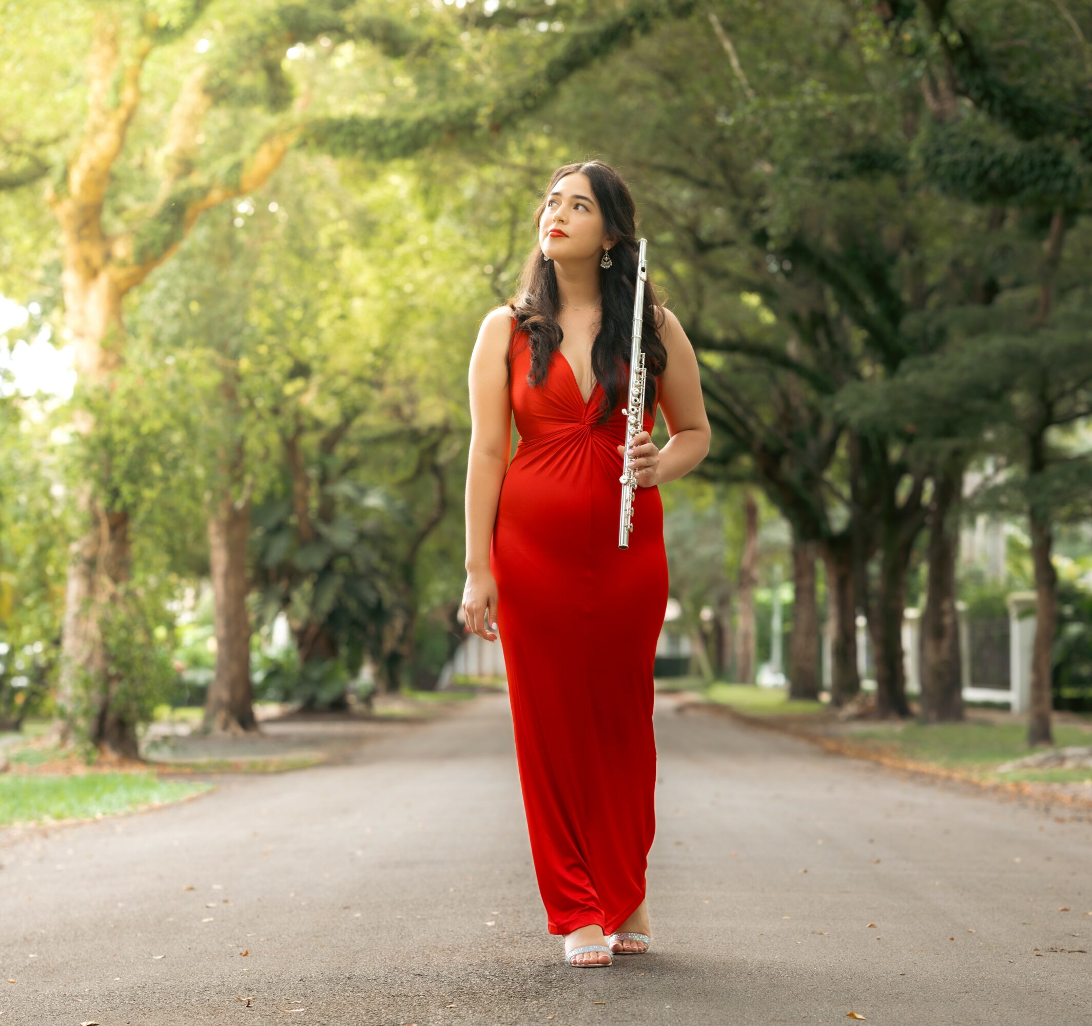 Woman in red dress walking on a tree-lined street holding a musical instrument.