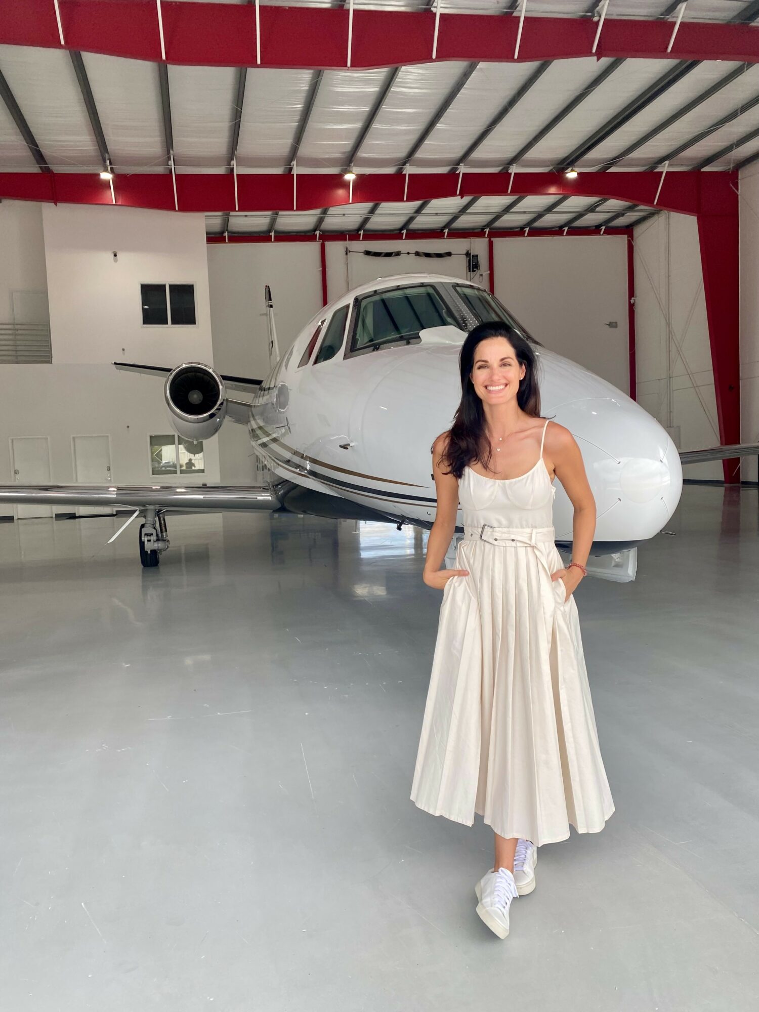 Woman in white dress standing in front of a private jet inside hangar.