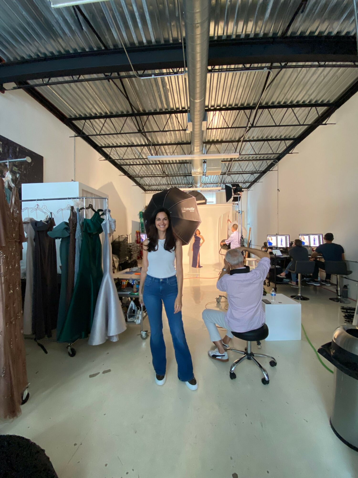 Woman standing in a room with clothing racks and a person sitting on a stool taking a photo.