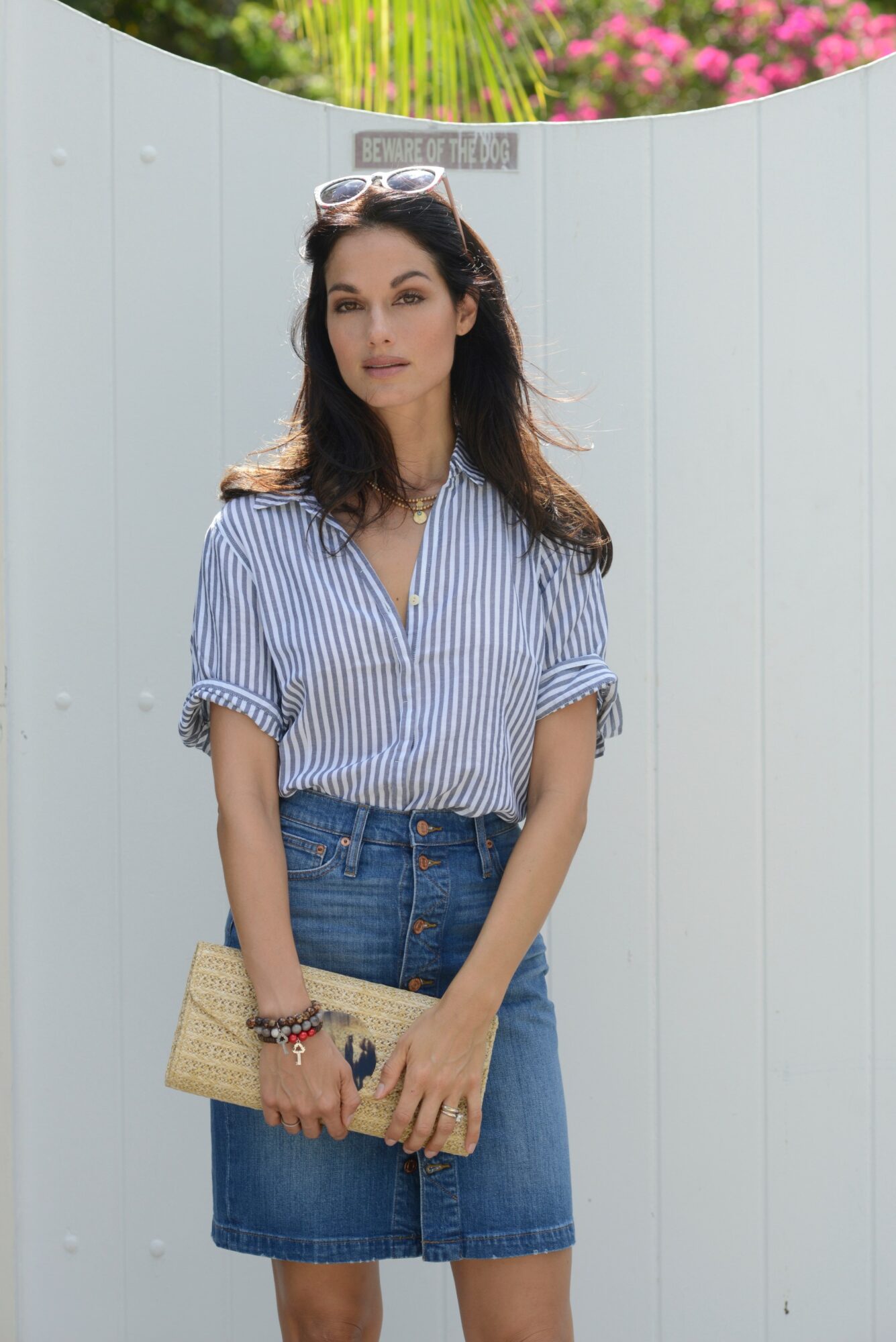 Woman with dark hair wearing sunglasses, striped shirt, and denim skirt holding a clutch bag outdoors.