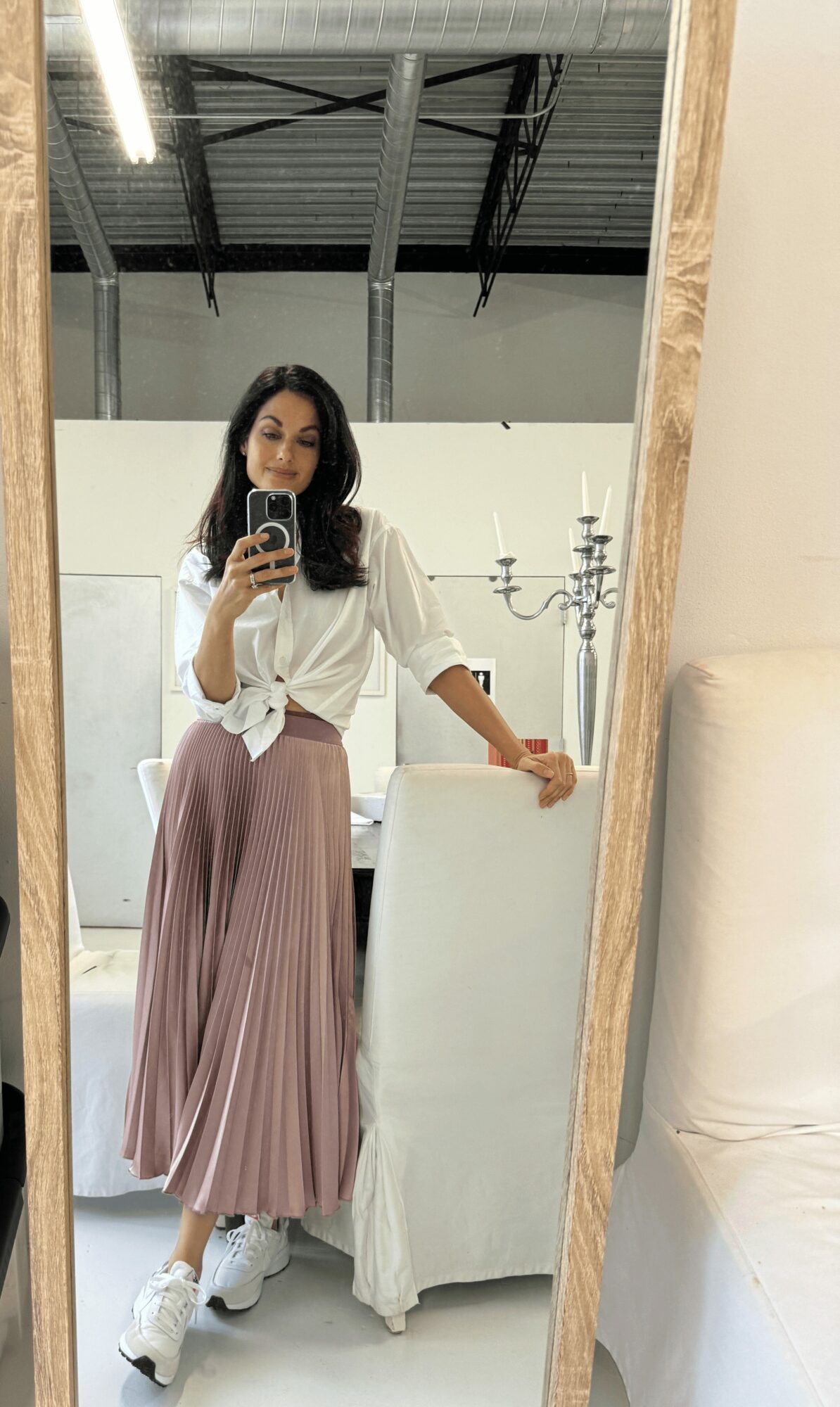 Woman taking a mirror selfie, wearing a white top and pink pleated skirt, standing in a room with chandelier and furniture.
