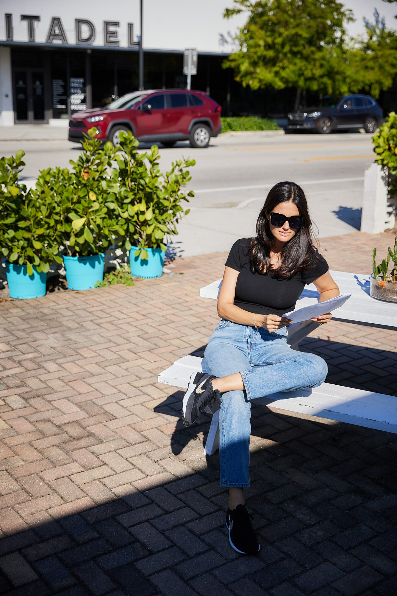 Woman sitting on a bench with sunglasses, black top, and jeans, reading a book outdoors on a sunny day.