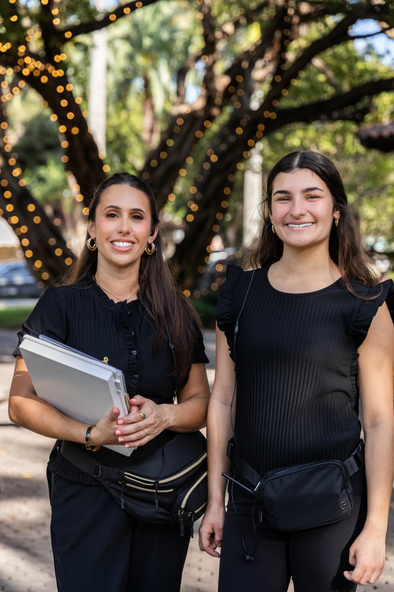 Two young women standing outdoors in front of a tree with string lights, smiling and holding notebooks.