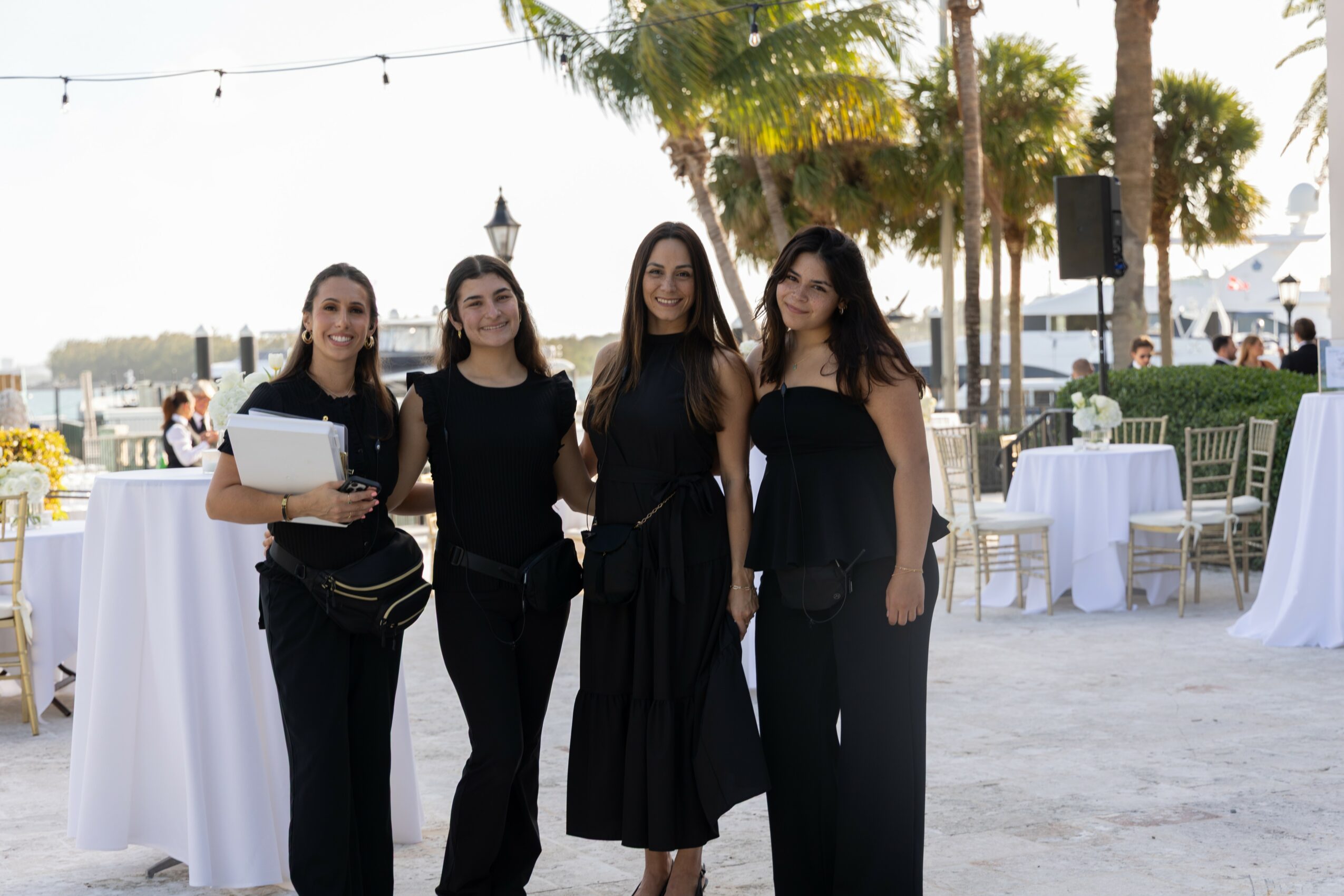 Four women standing outdoors near tables and palm trees, smiling at the camera.