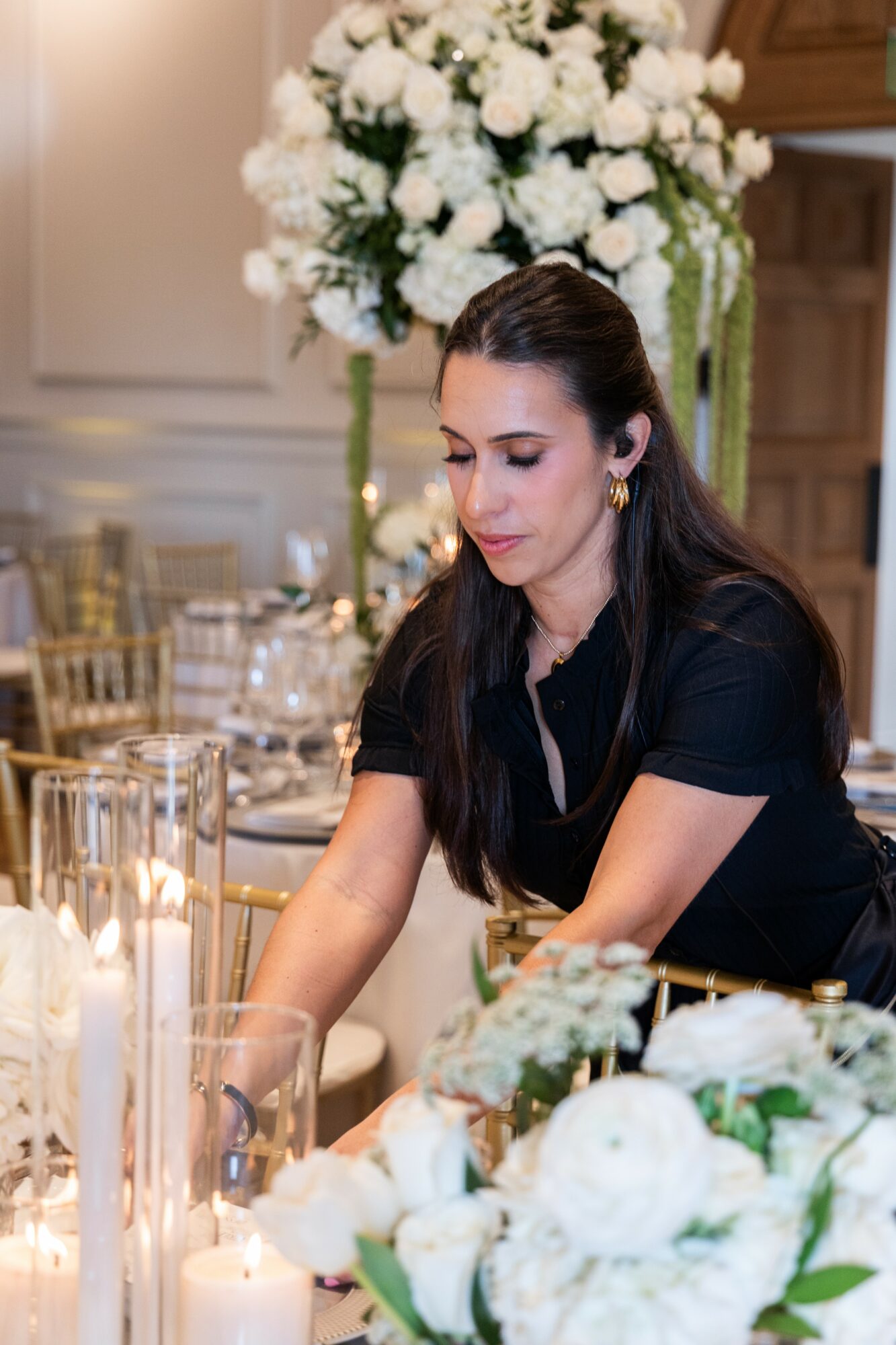 Woman with dark hair and earrings seated at a table with floral arrangements and candles.
