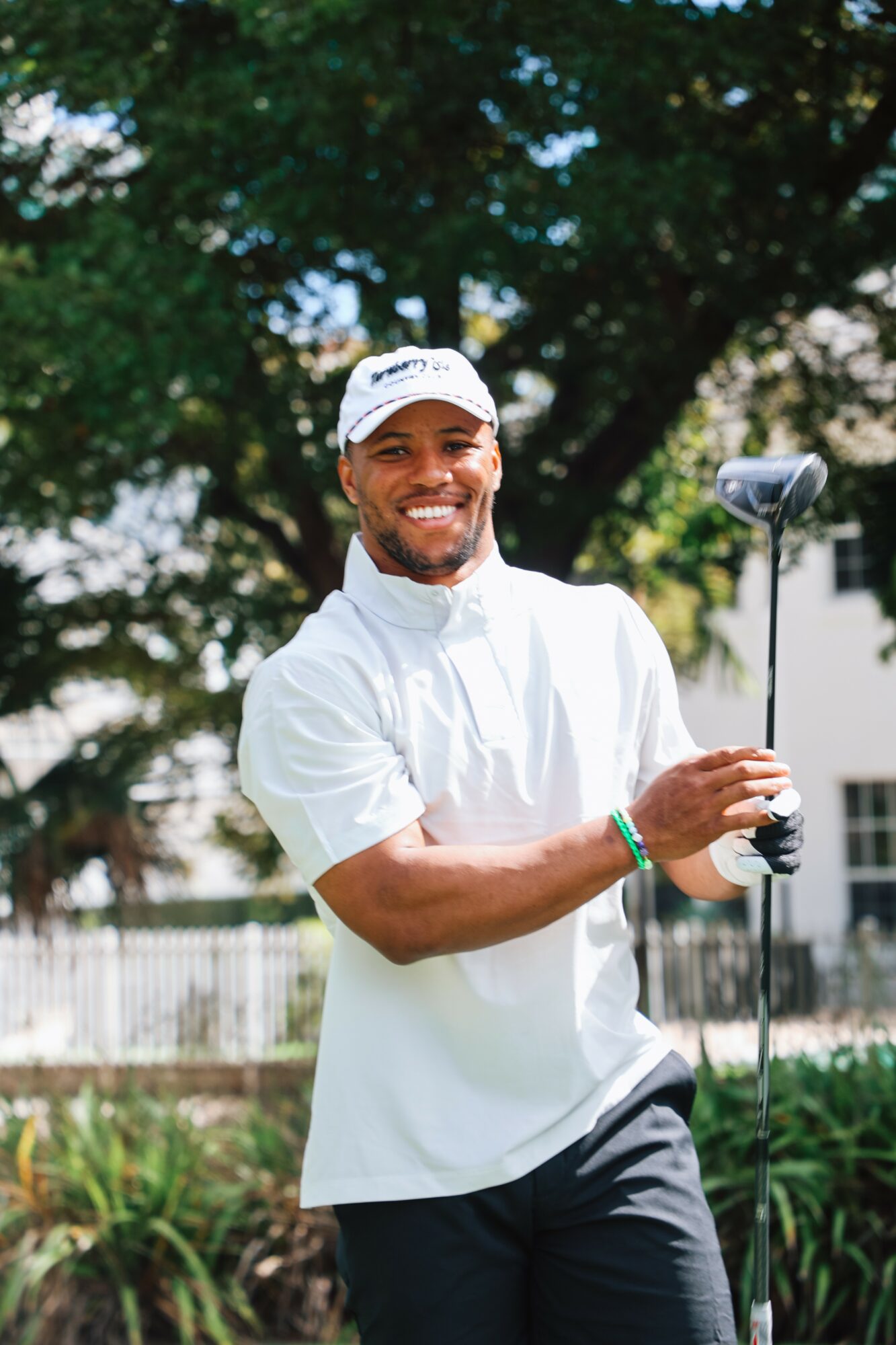 Man smiling outdoors holding a golf club, wearing a white shirt and cap, with trees in background.