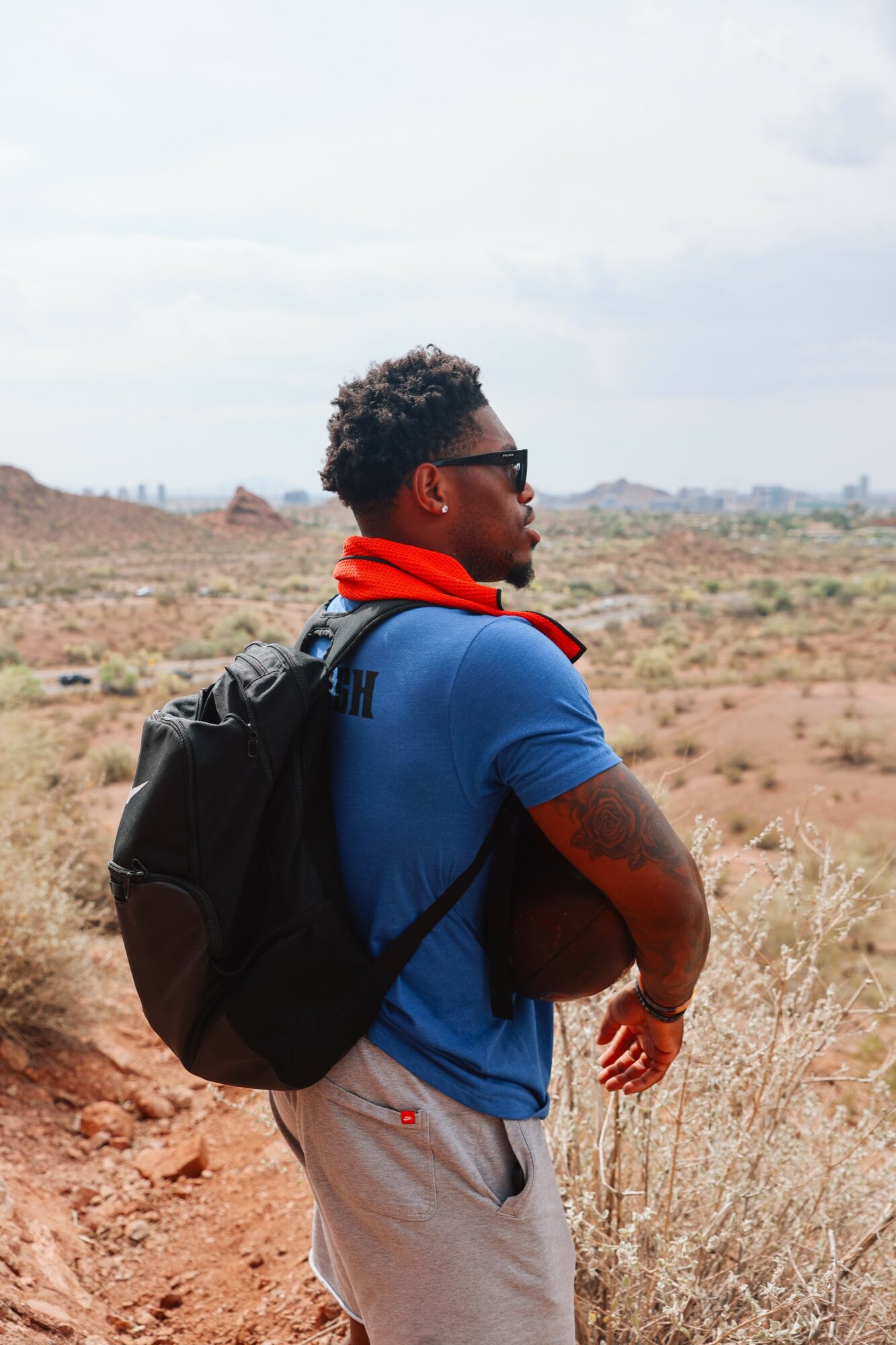 Young man with sunglasses and backpack standing in a desert landscape, holding a ball, facing sideways.