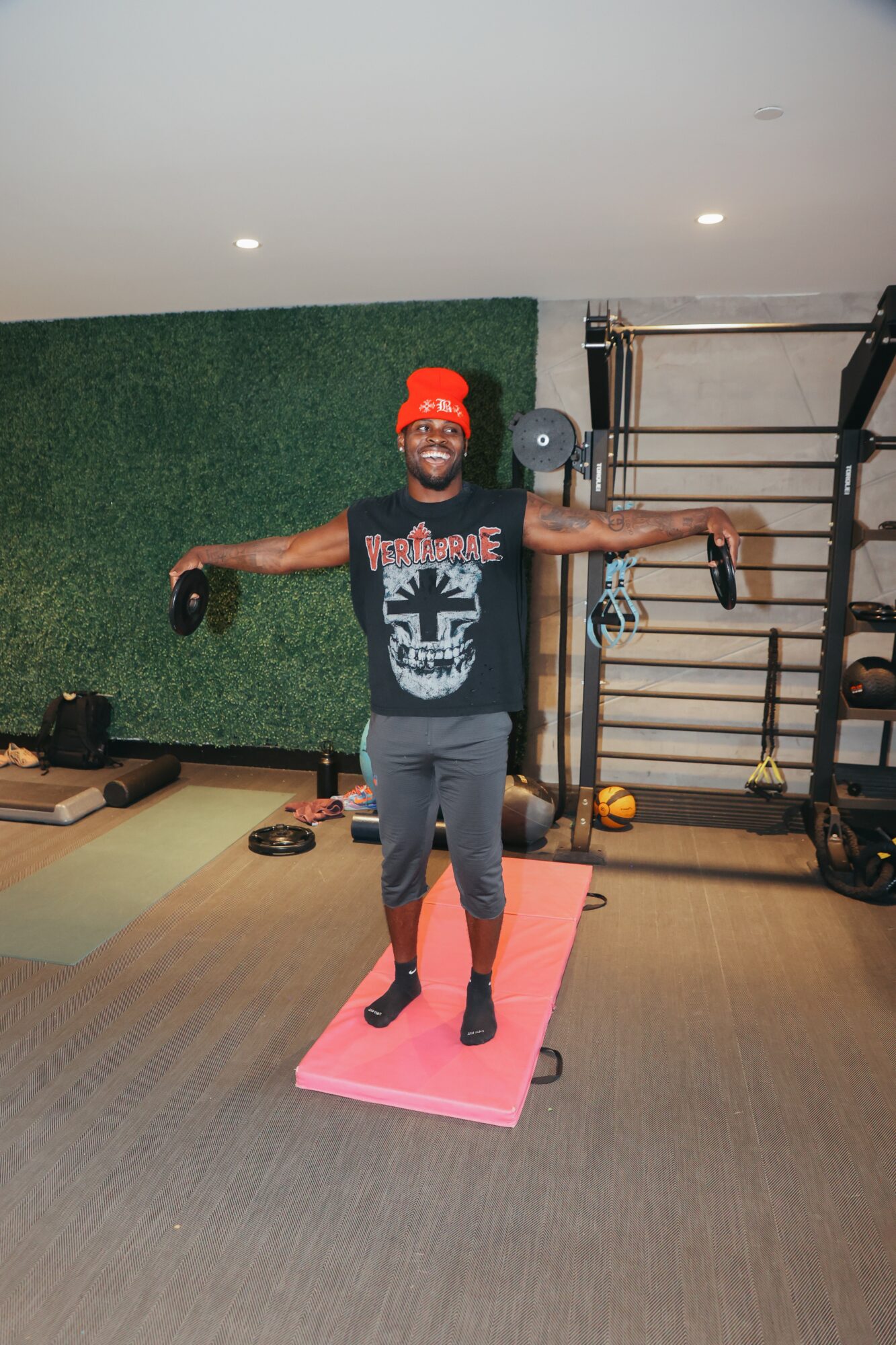 Person standing on a pink exercise mat with arms outstretched in a gym, wearing a red beanie and black t-shirt.