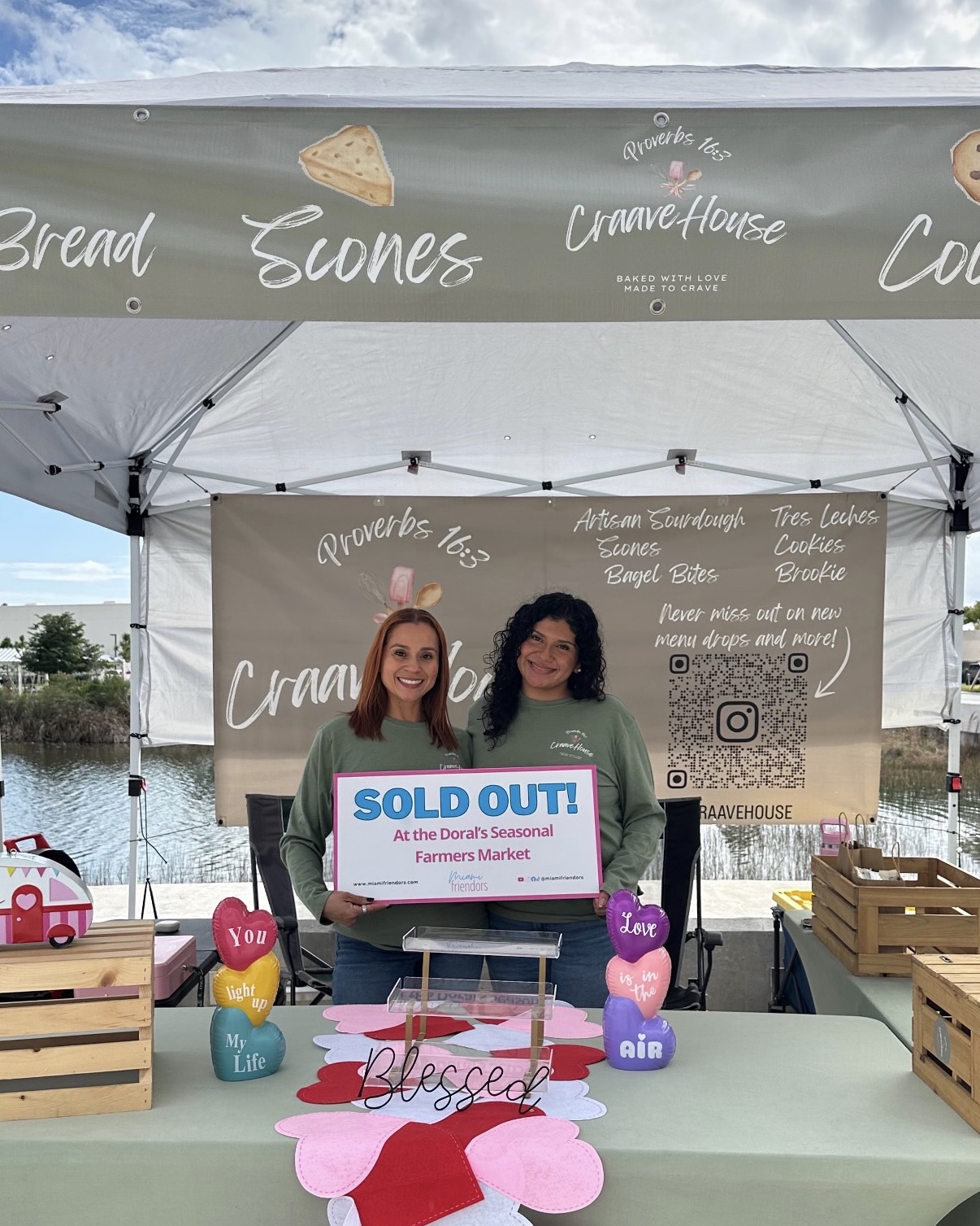 Two women holding a sold out sign in front of a market stall with bread and scones banners, colorful heart-shaped balloons, and wooden crates.