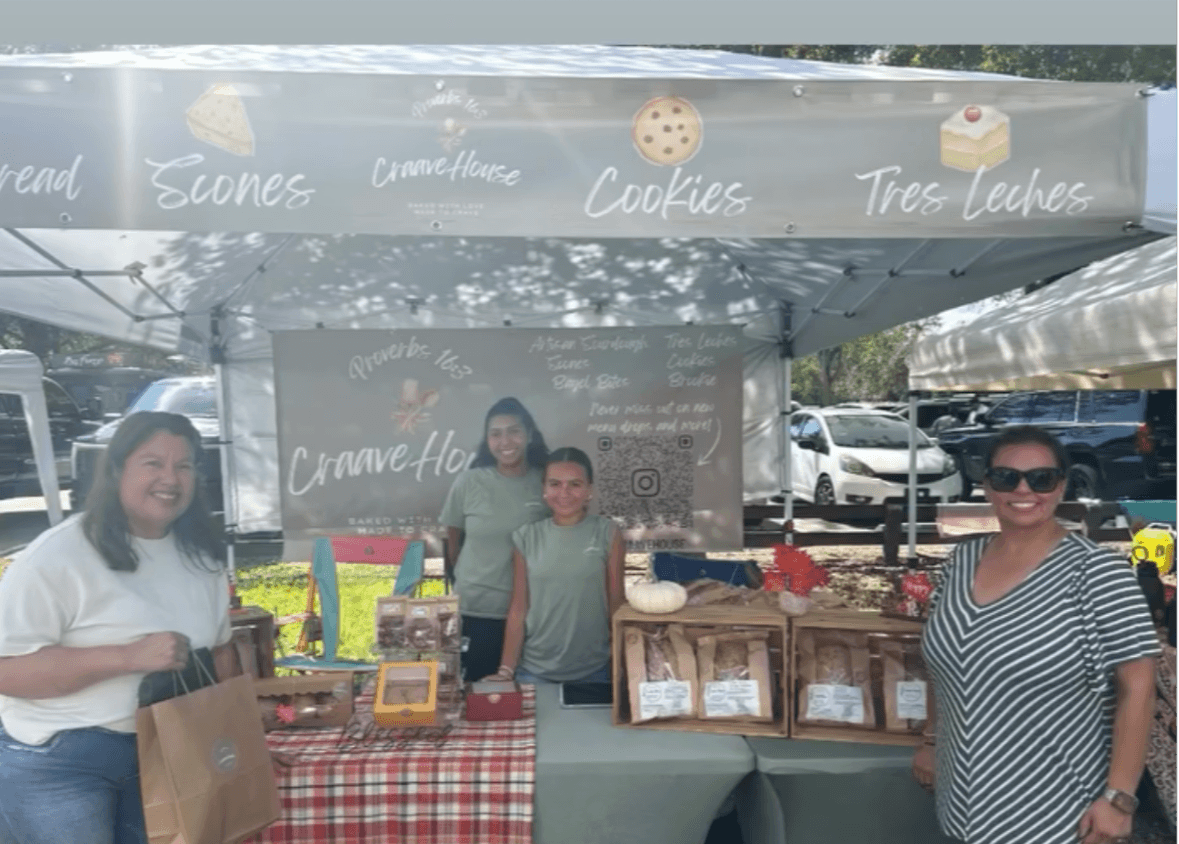 Three women stand at a cookie stand with a gray banner and baked goods in bags on a table.