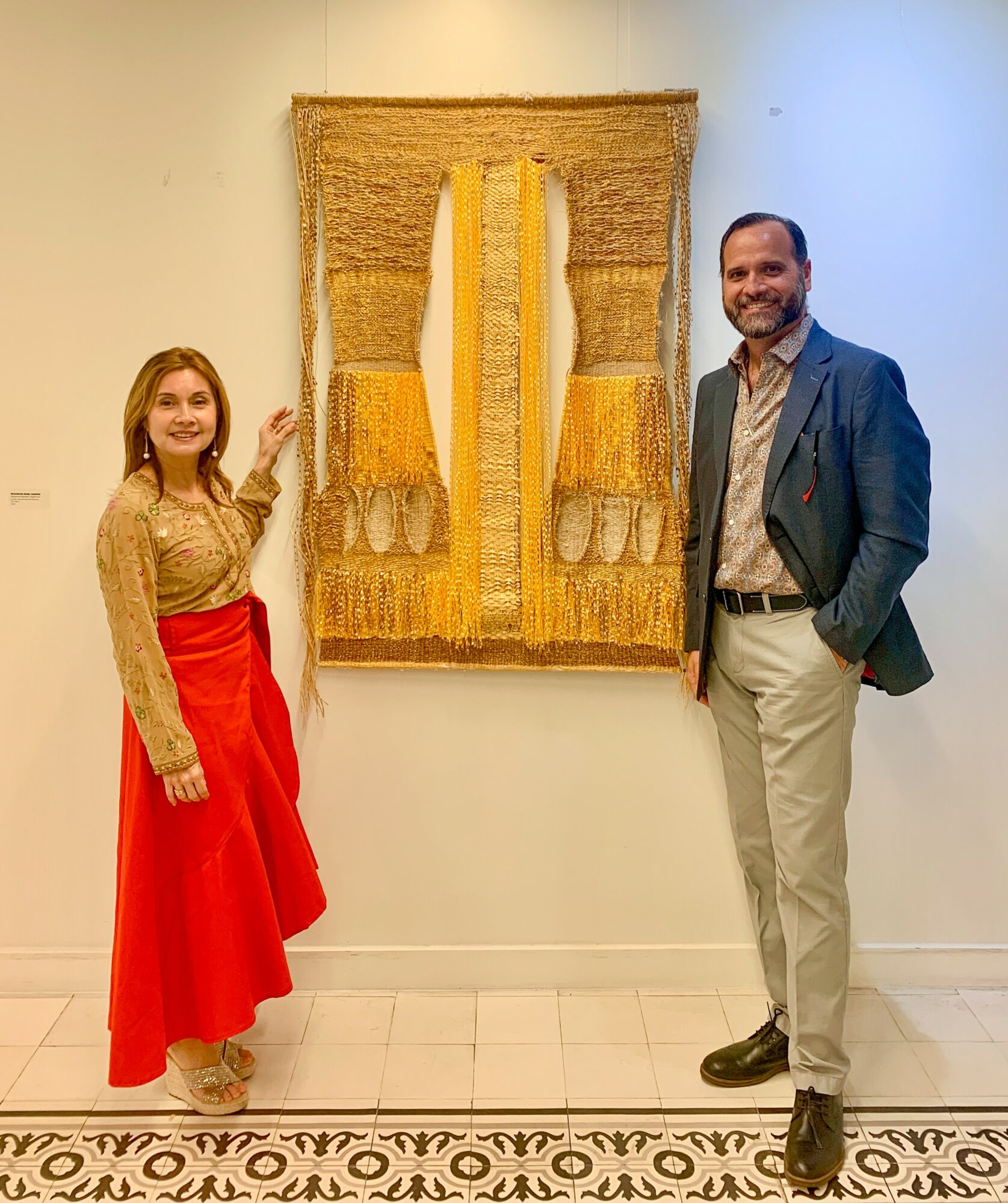 Two people stand beside a textured wall hanging with yellow and beige tones, in an indoor gallery setting.