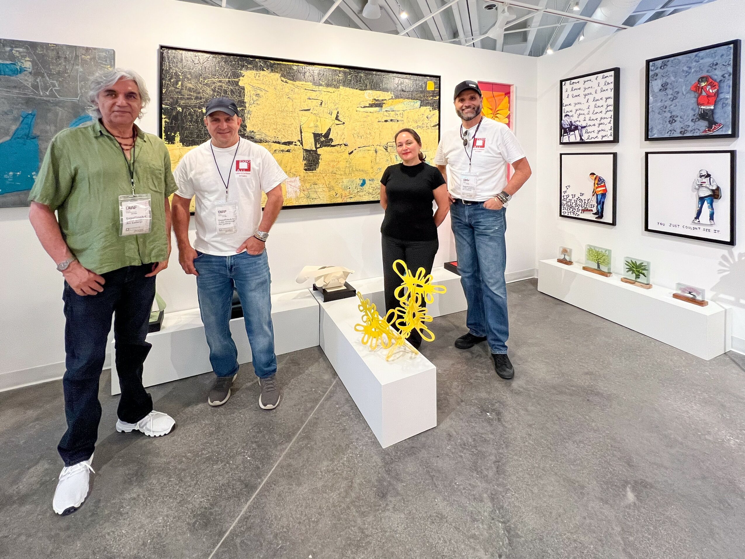 Four people stand in an art gallery with paintings on white walls, some sitting on white benches, gray carpeted floor.