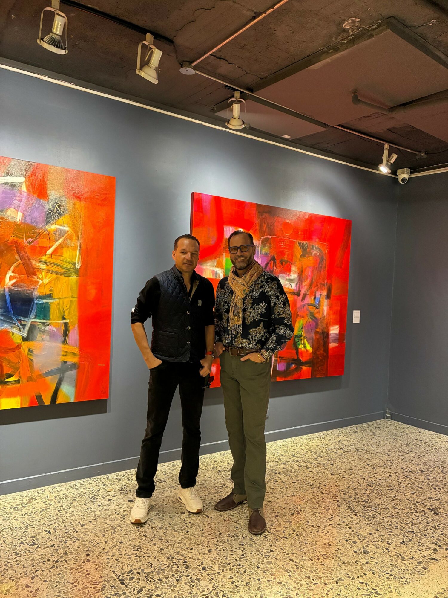 Two men stand in front of colorful abstract paintings on a gray gallery wall.