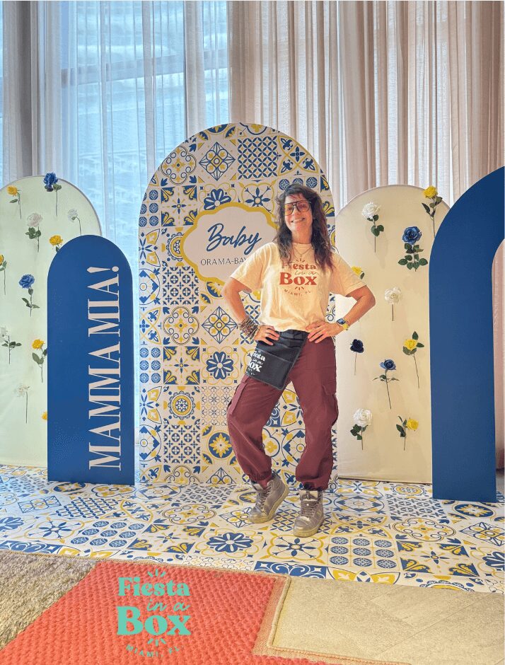 Woman standing in front of decorative backdrop with floral and geometric patterns, and signs reading 'MAMMAMIA' and 'Felicity Box'.