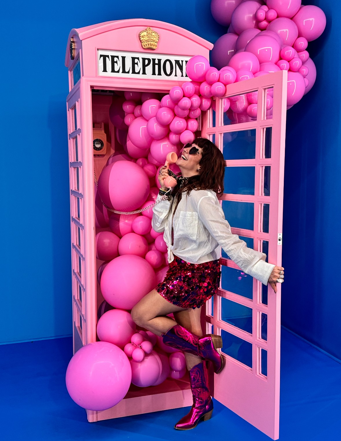 Woman in sunglasses and floral skirt poses inside a pink telephone booth decorated with pink balloons against a blue background.