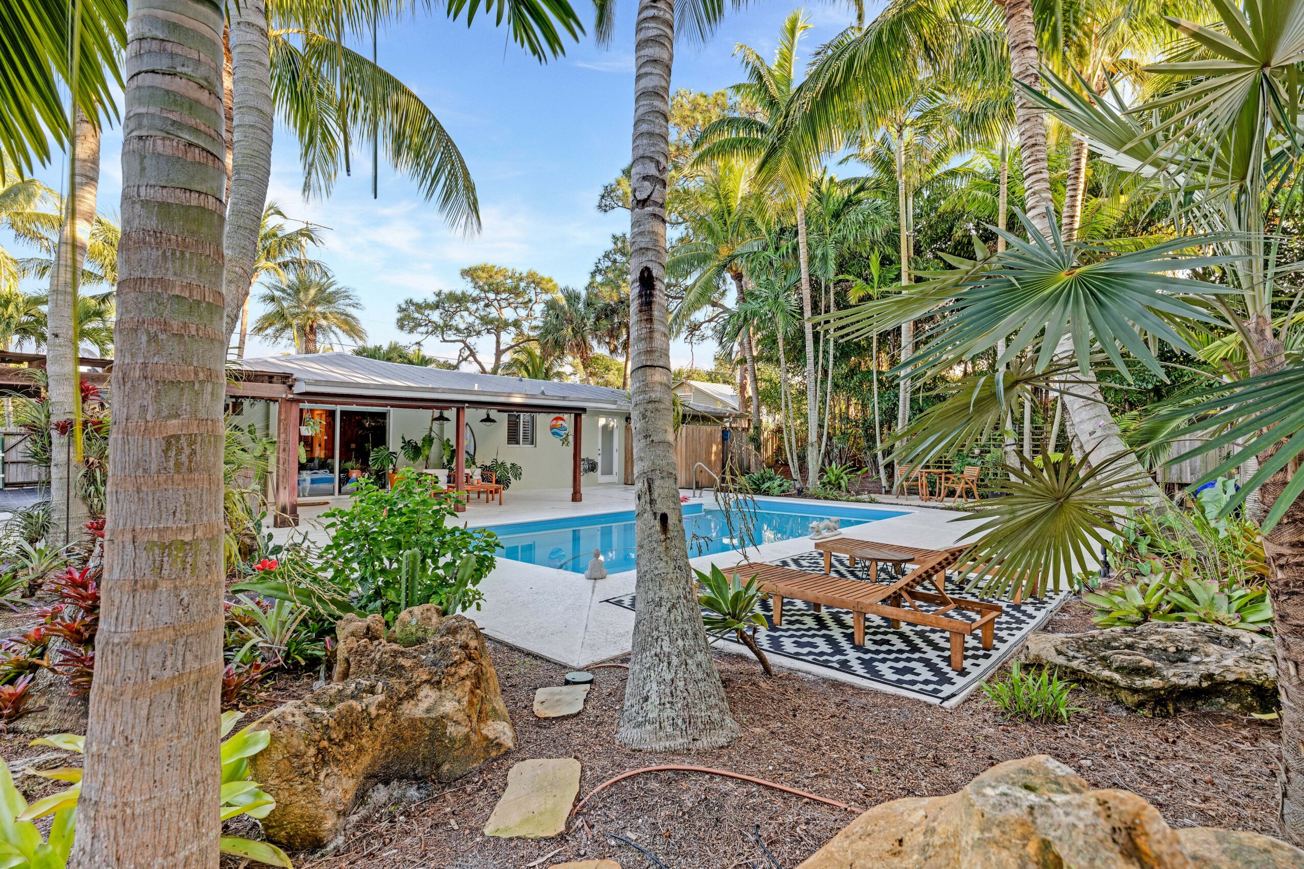 Pool area surrounded by palm trees and tropical plants, with lounge chairs and a patterned outdoor rug.