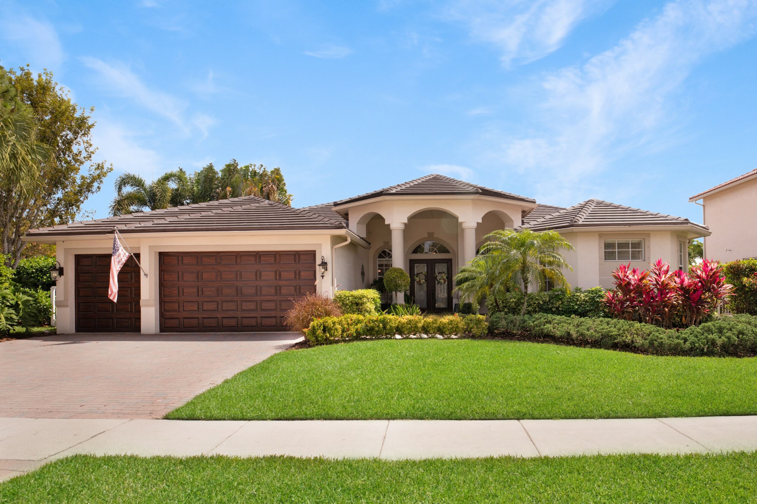 Single-story house with a two-car garage, front yard with green lawn, bushes, and flowering plants, under a blue sky.