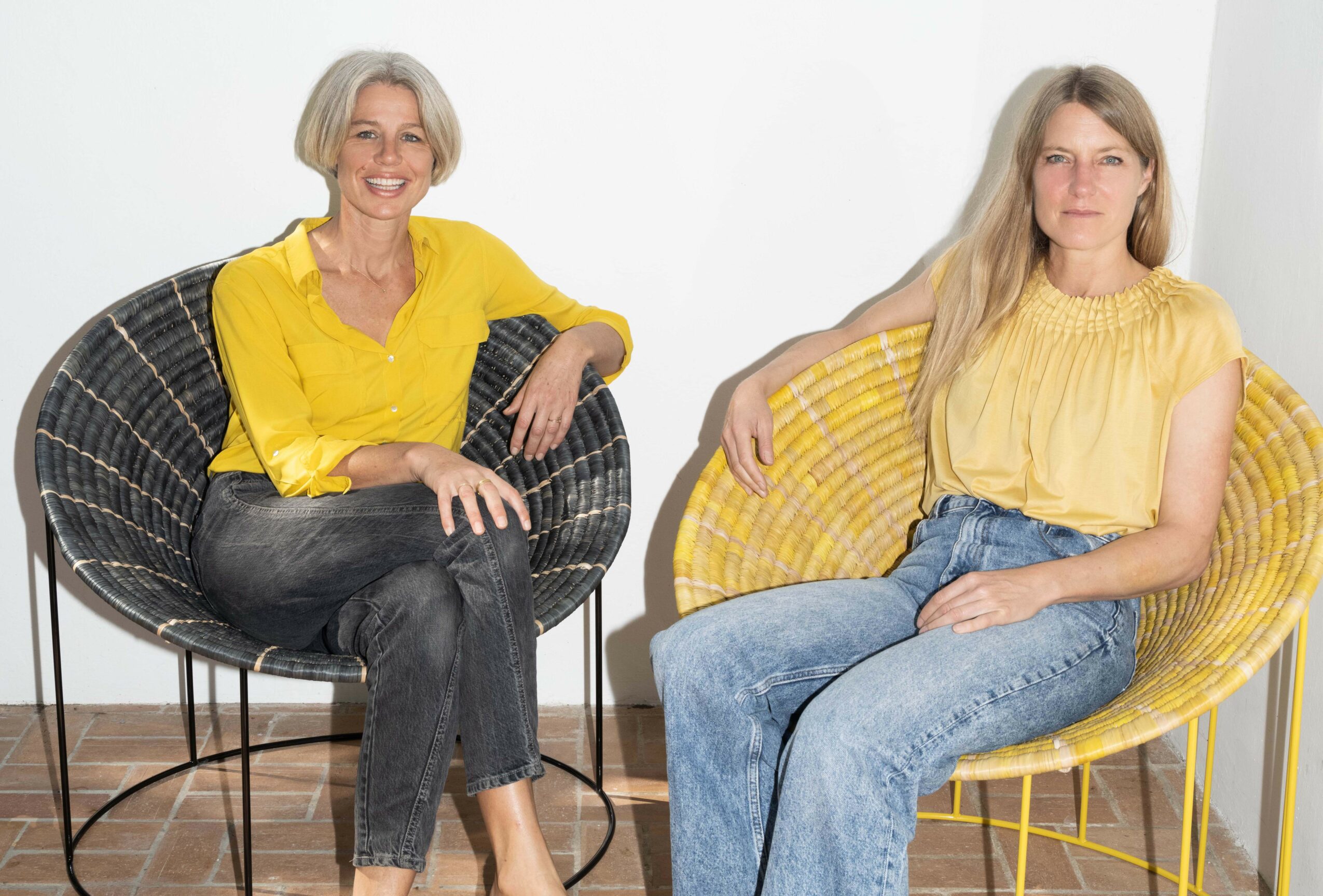 Two women sitting in yellow chairs, smiling, against a white wall, wearing yellow tops and jeans.