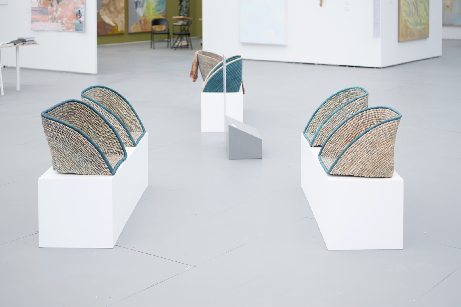 Six woven chairs on white pedestals arranged in a semi-circle in an art gallery.