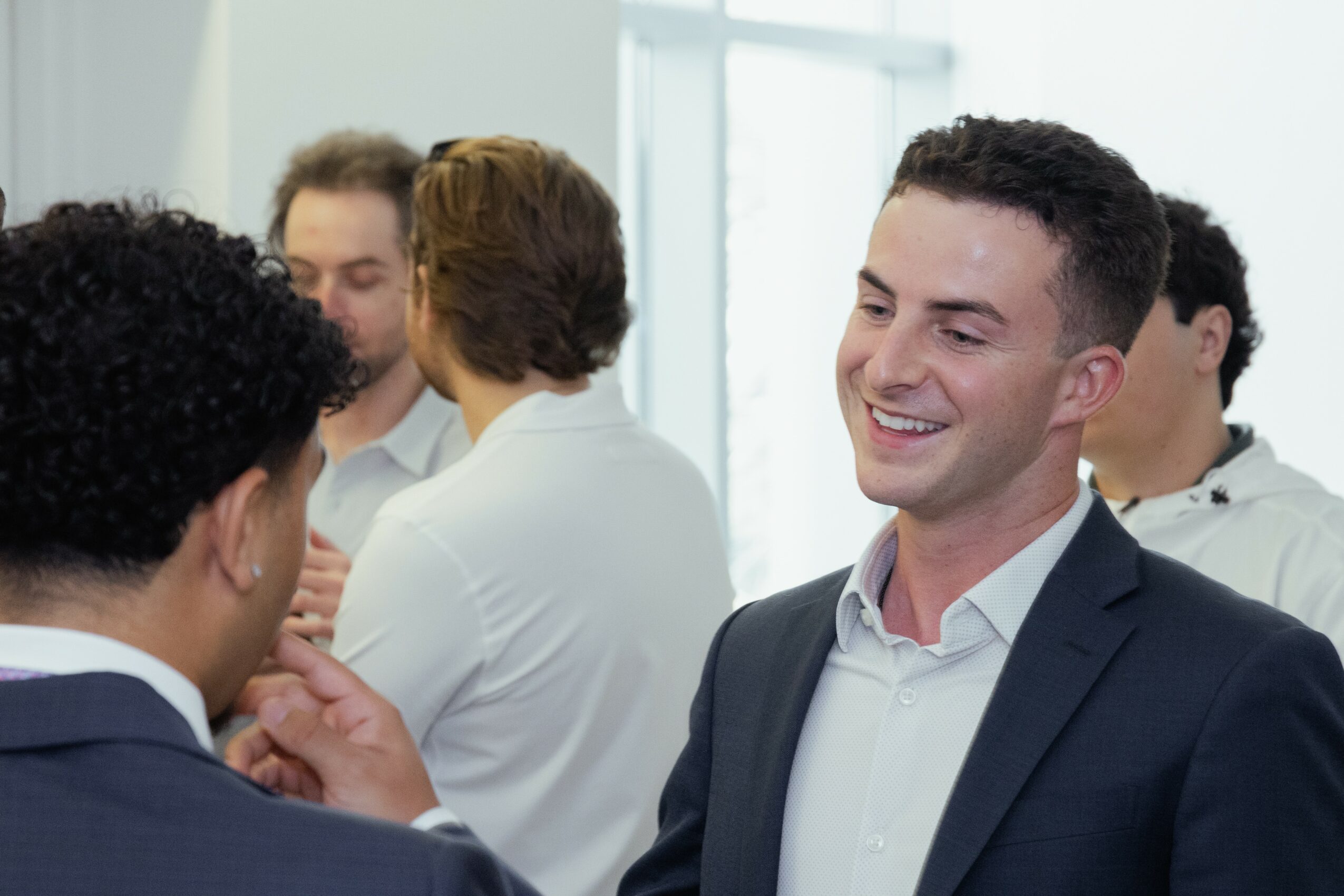 Group of people talking and smiling indoors, some facing each other, in a bright room with large windows.