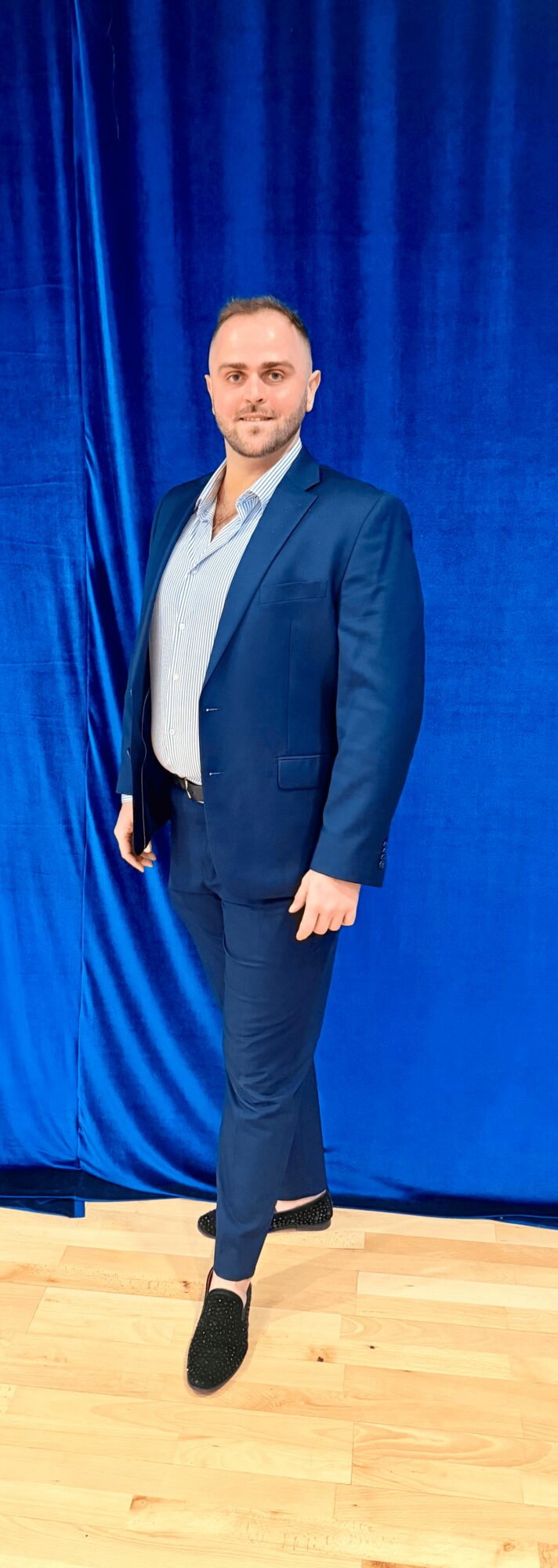 Man in a blue suit standing on a wooden floor in front of a blue curtain.