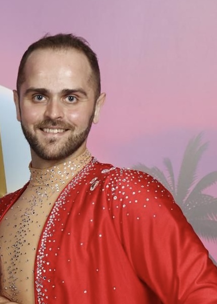 Man with short hair and beard smiling, wearing a red costume with rhinestones, background with pink sky and palm trees