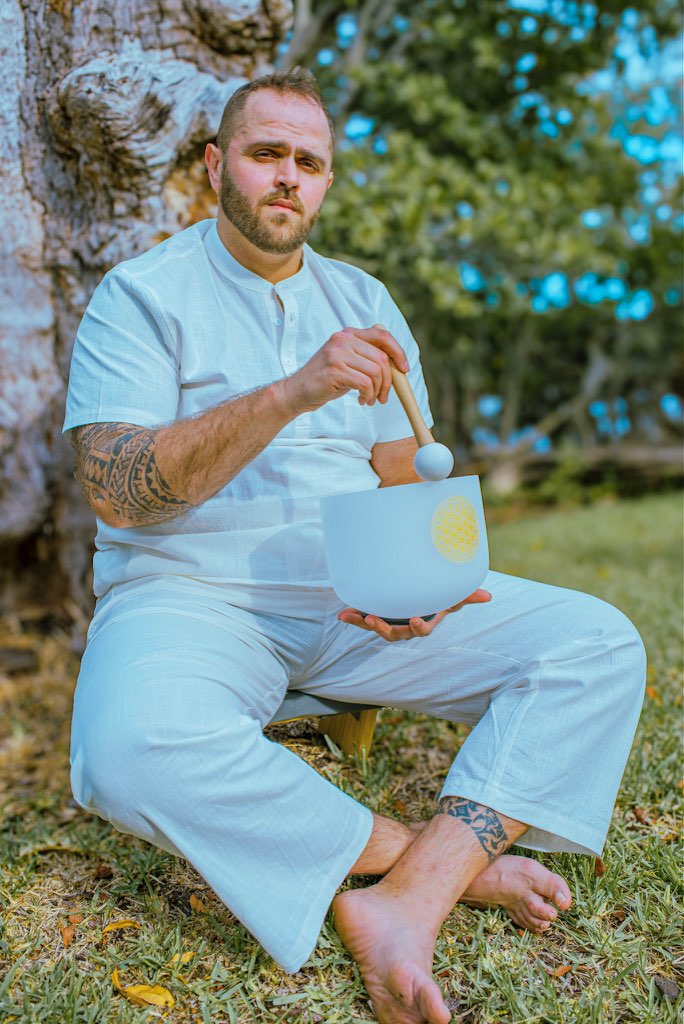 Man sitting outdoors on grass, holding a singing bowl and mallet, with trees and blue sky in background.