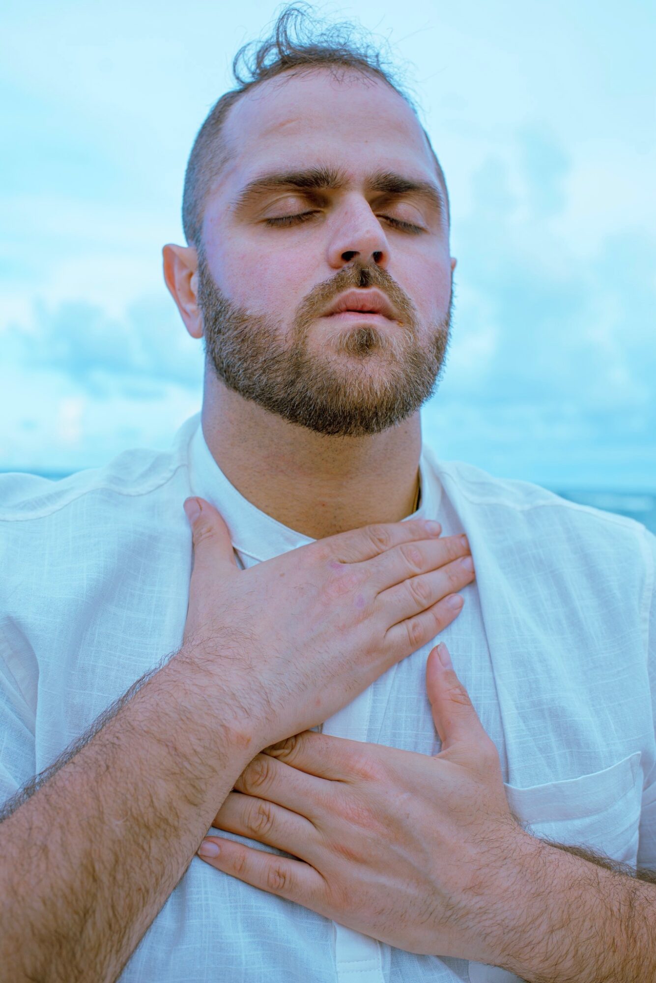 Man with eyes closed, hands on chest, outdoors with cloudy sky background.