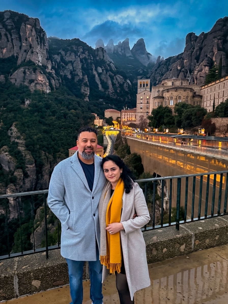 Two people stand on a bridge with mountains, a river, and historic buildings in the background during evening.