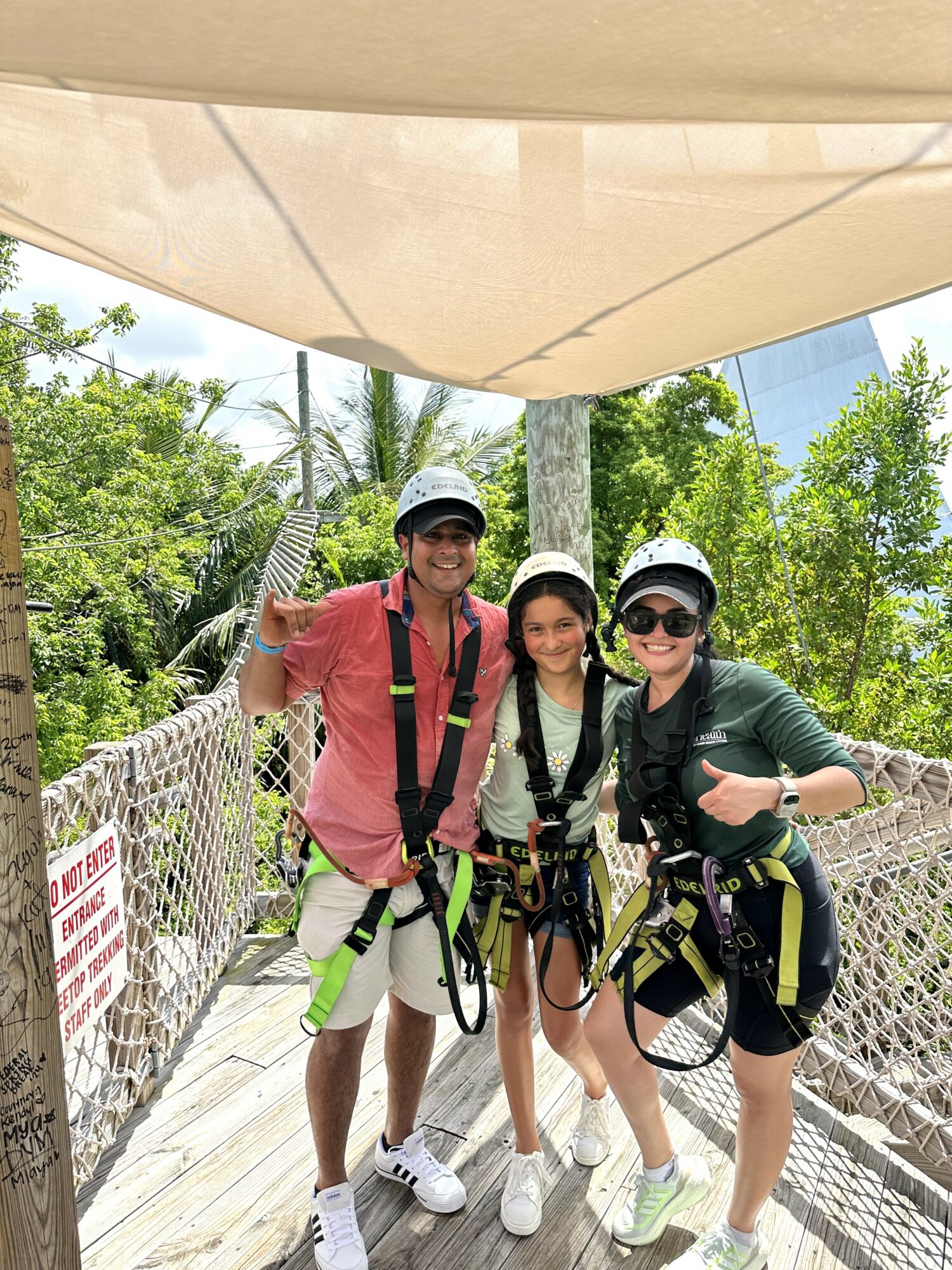 Three people in harnesses and helmets stand on a wooden bridge outdoors, smiling, with trees and a building in background.